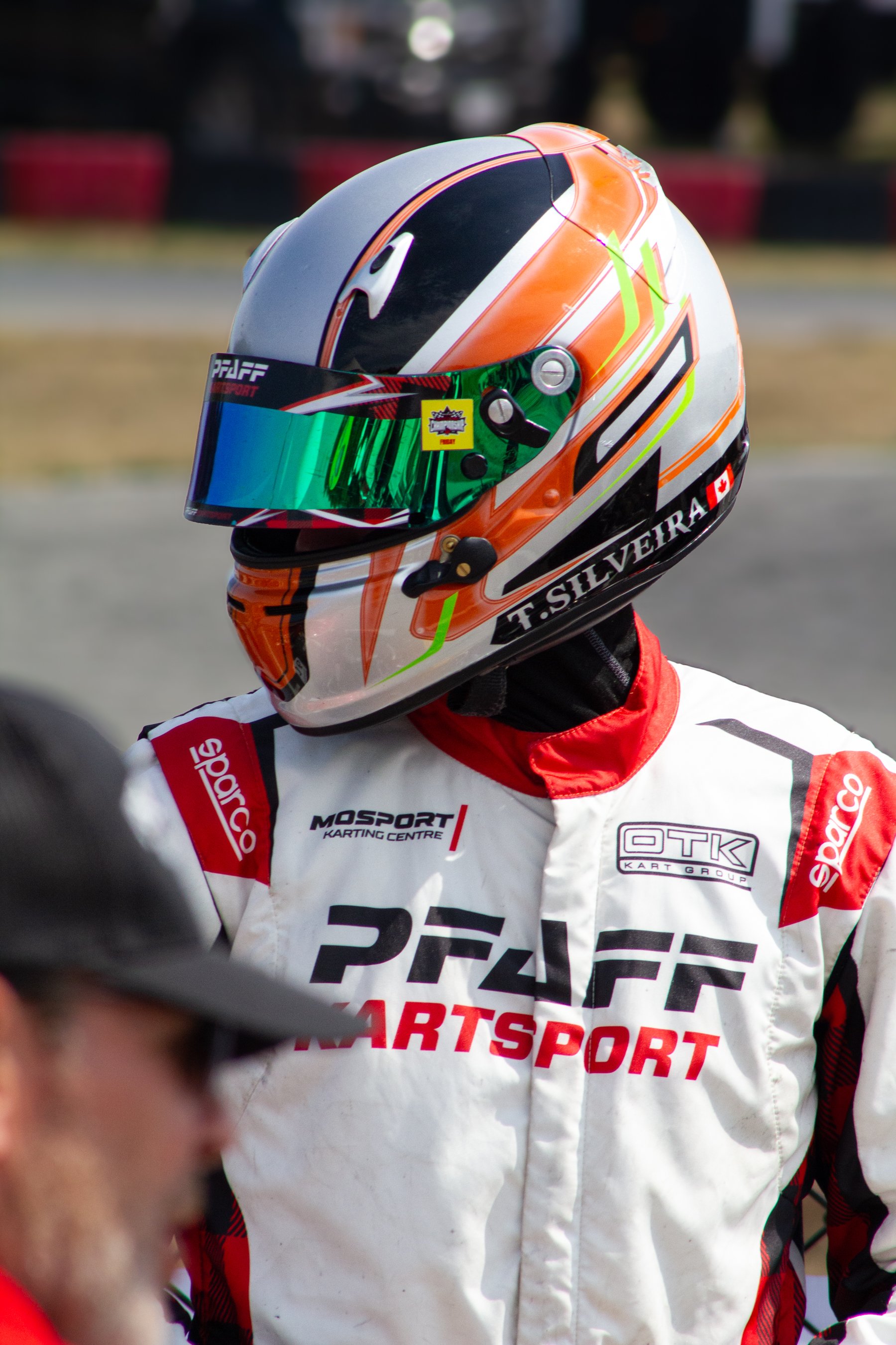 Race car driver wearing a colorful helmet and a white racing suit with red and black accents, with various sponsor logos including PFAFF, MOSPORT, and OTG, standing on a racetrack.