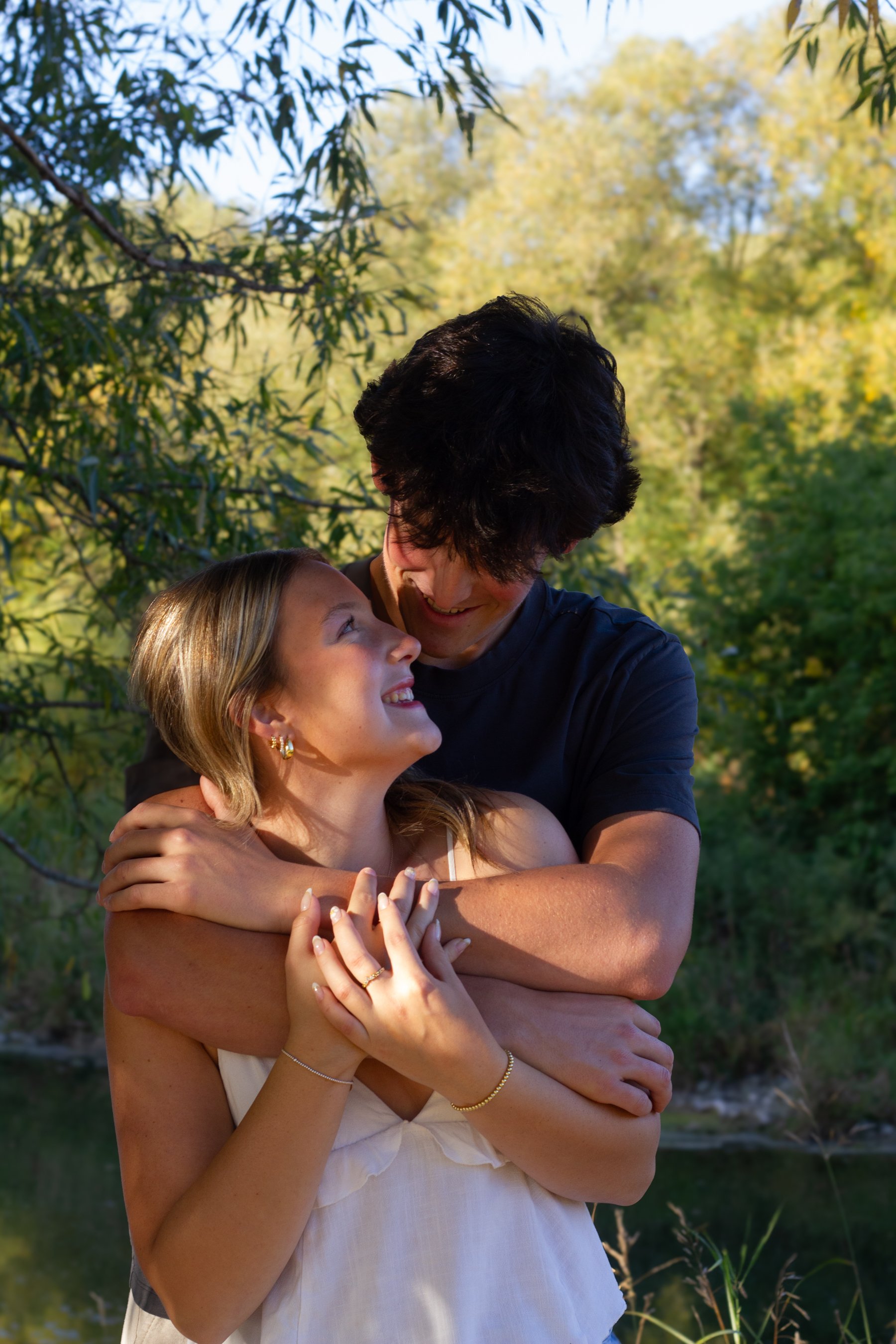 A couple is embracing outdoors near a river, with trees in the background. The woman is smiling up at the man, who is holding her in a hug, both appearing happy and enjoying each other's company.