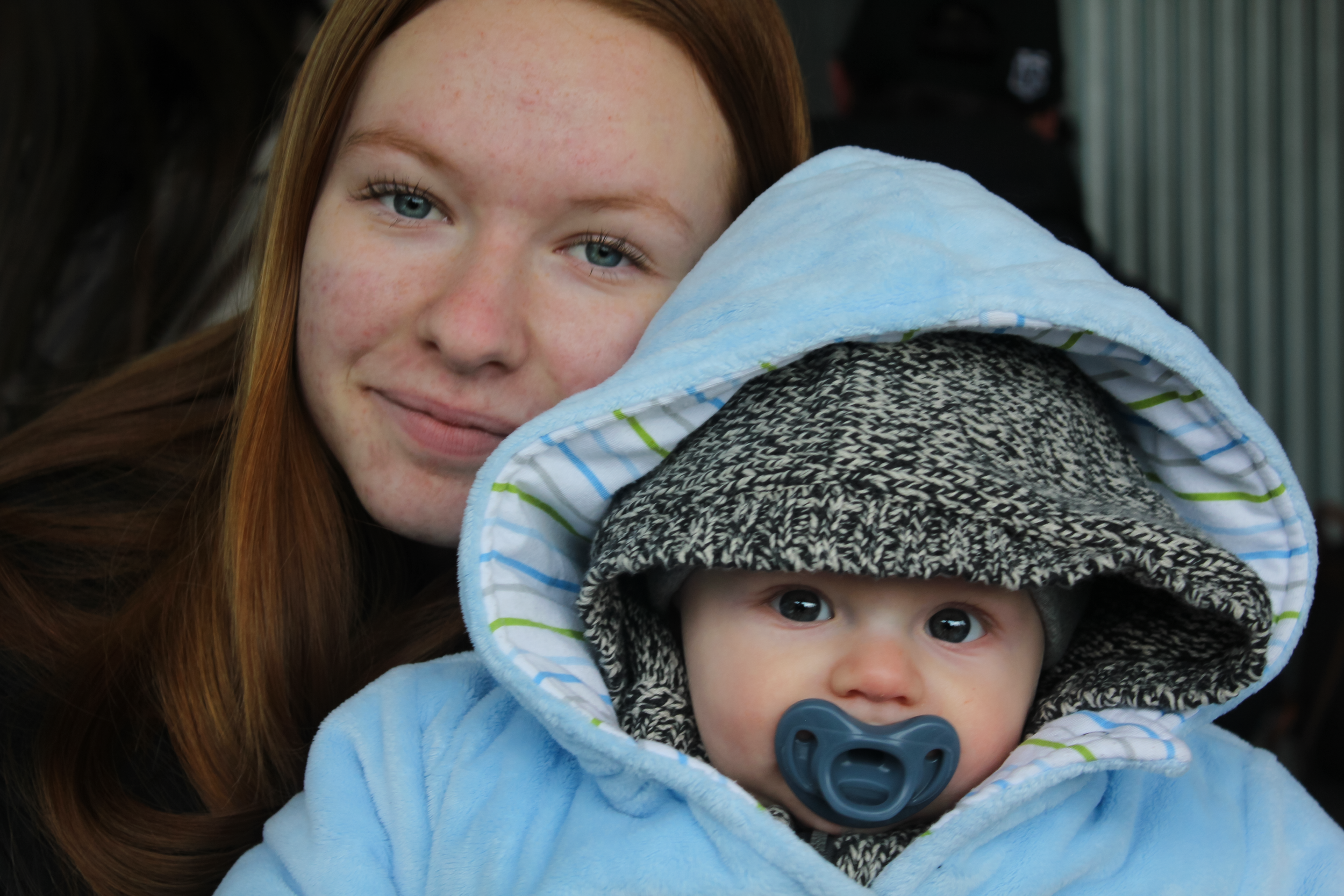 A woman with long red hair and blue eyes smiling next to a baby with a pacifier and wearing a hooded coat with a hat inside.