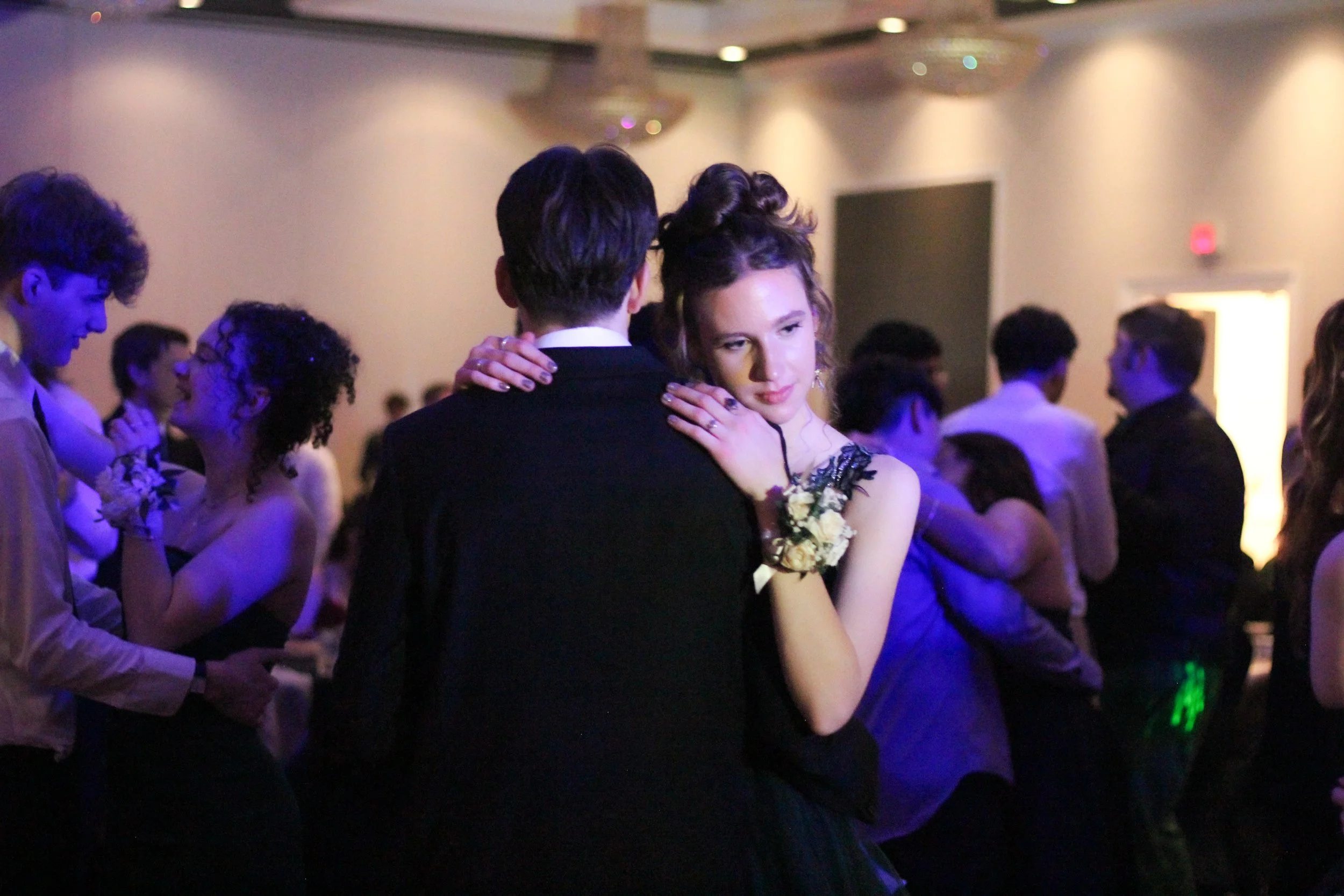 Couple dancing closely at a formal dance or prom, with other couples dancing in the background, indoors with dim lighting.