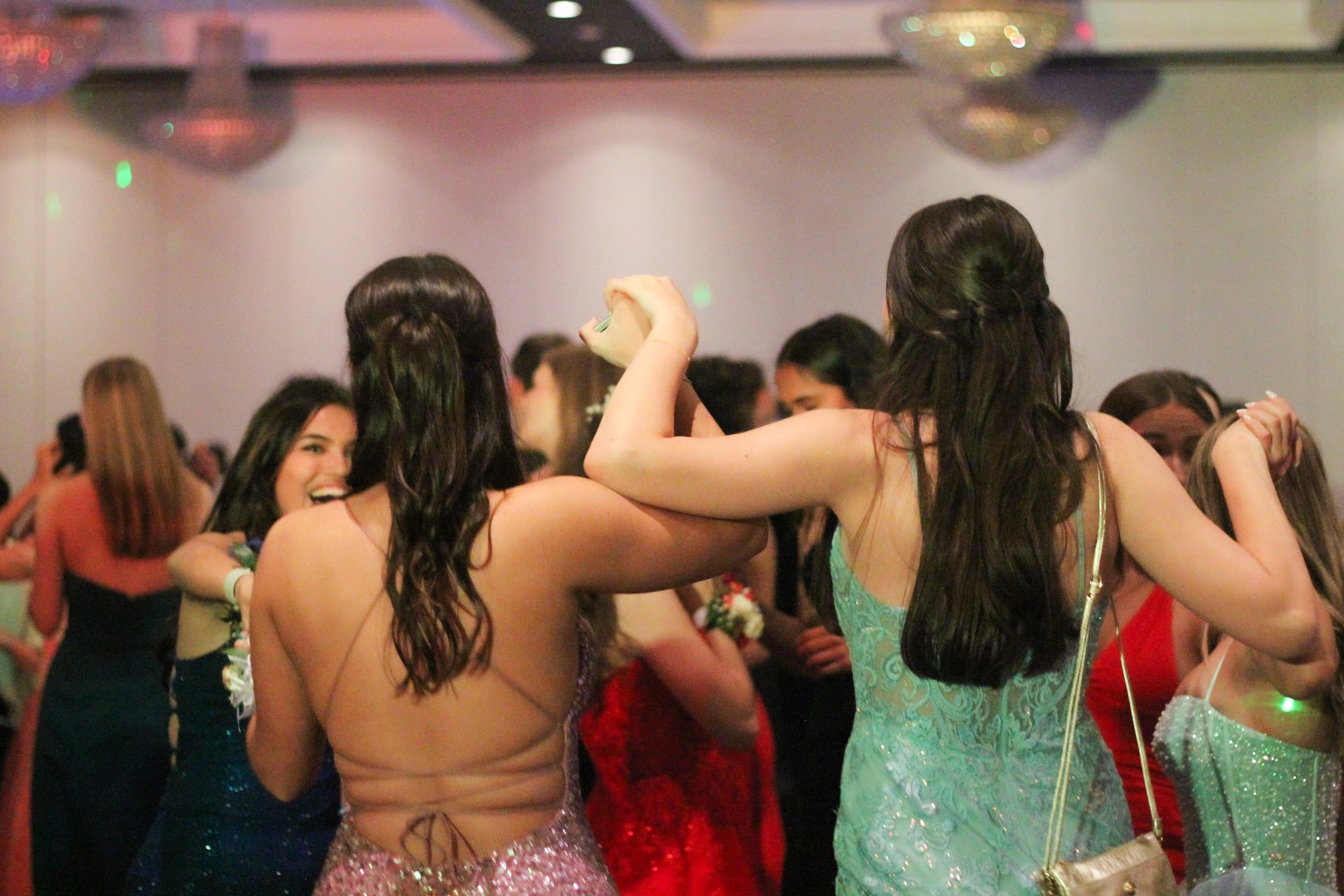Two young women in formal dresses, one wearing a pink dress and the other wearing a blue dress, are dancing and holding hands at a party or dance event, with other people in the background.