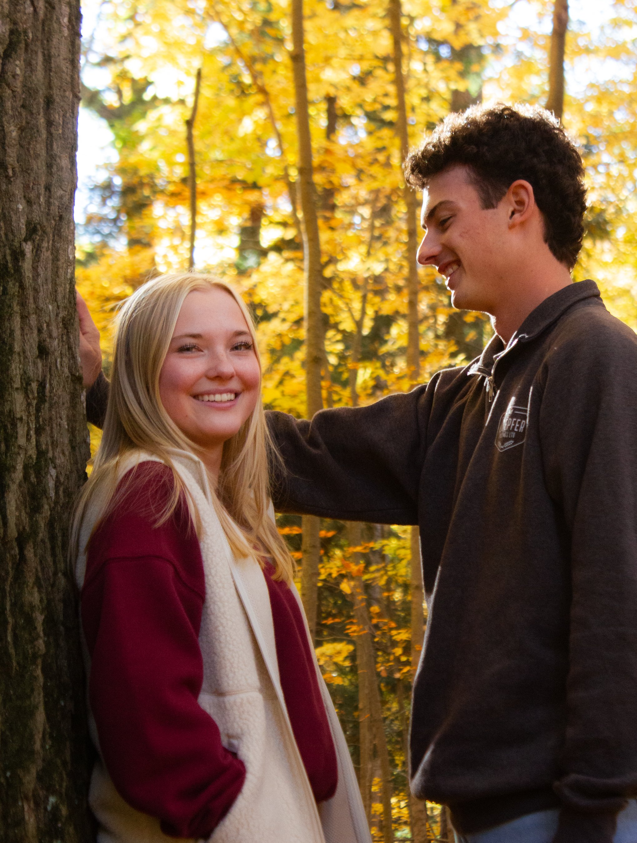 A young man and woman smiling in a forest with yellow autumn leaves. The man has curly dark hair and is wearing a black hoodie, while the woman has long blonde hair and is wearing a white and red jacket.