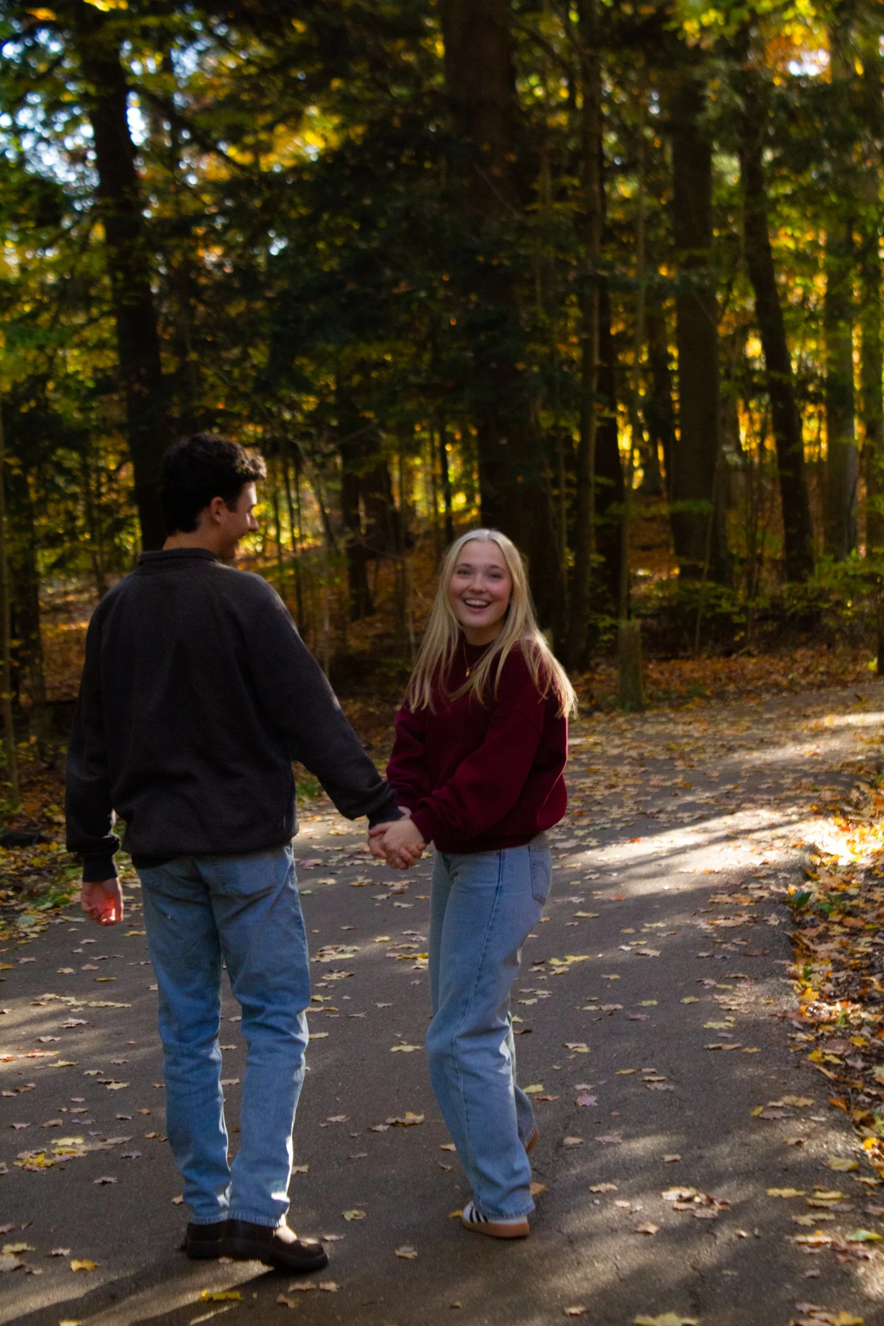 A young man and woman holding hands and smiling on a wooded trail in autumn.