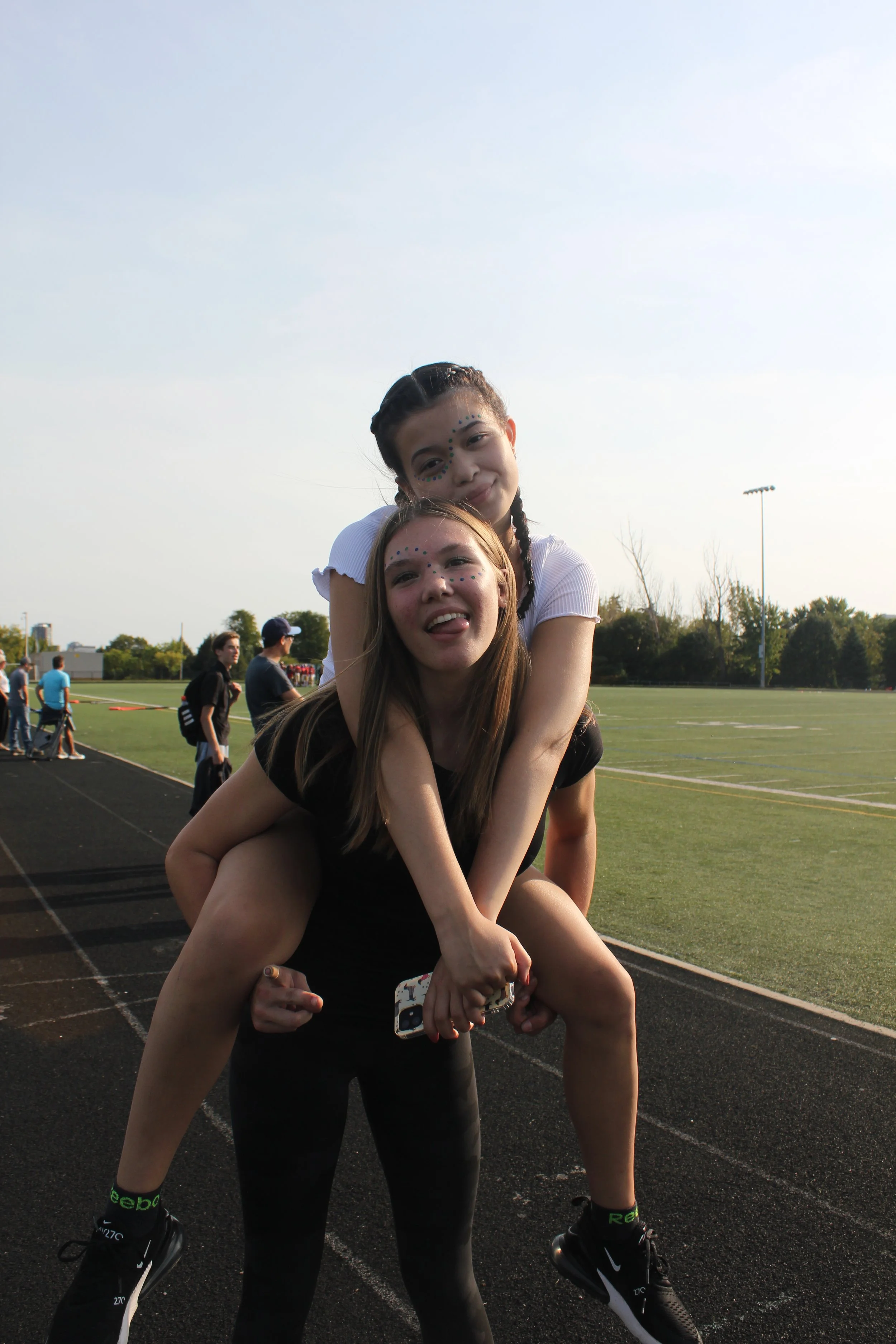 Two young women, one giving a piggyback ride to the other, smiling and making faces on a track field during daytime with a grassy field and other people in the background.