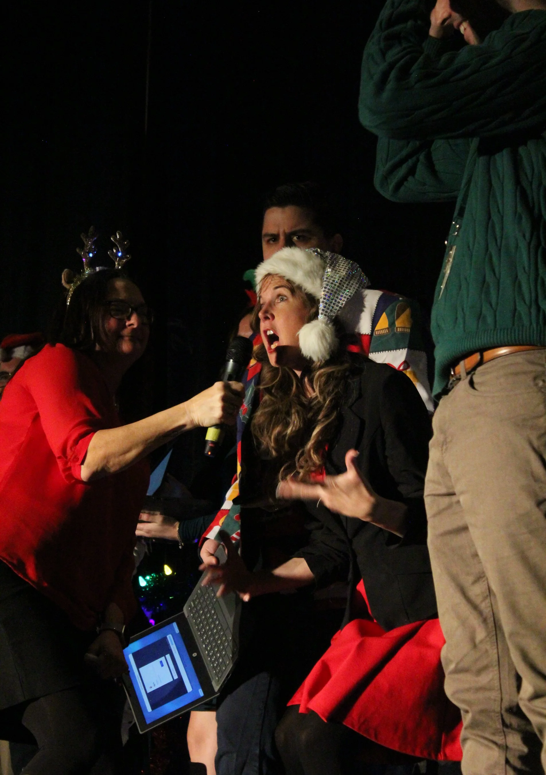 Group of people at a Christmas party, dressed in holiday attire, with one woman wearing a Santa hat and reindeer antlers, speaking into a microphone, while others hold a laptop and stand around her.