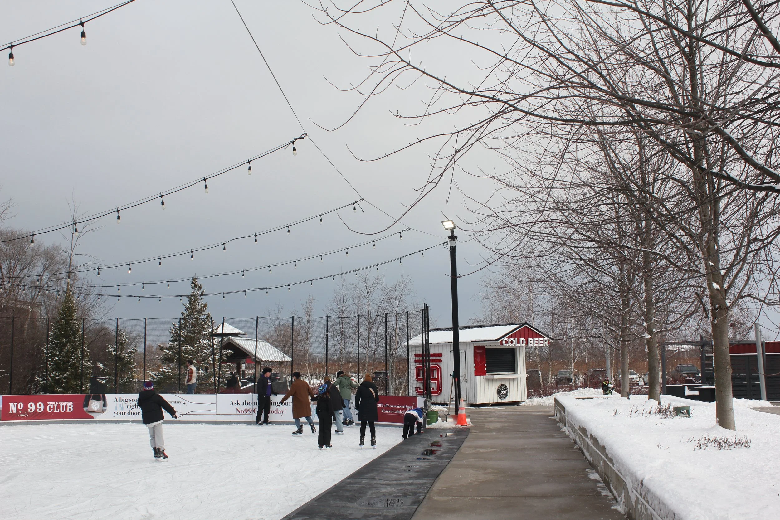 People ice skating at an outdoor rink surrounded by snow-covered trees and a sign for cold beer, under string lights on a cloudy winter day.