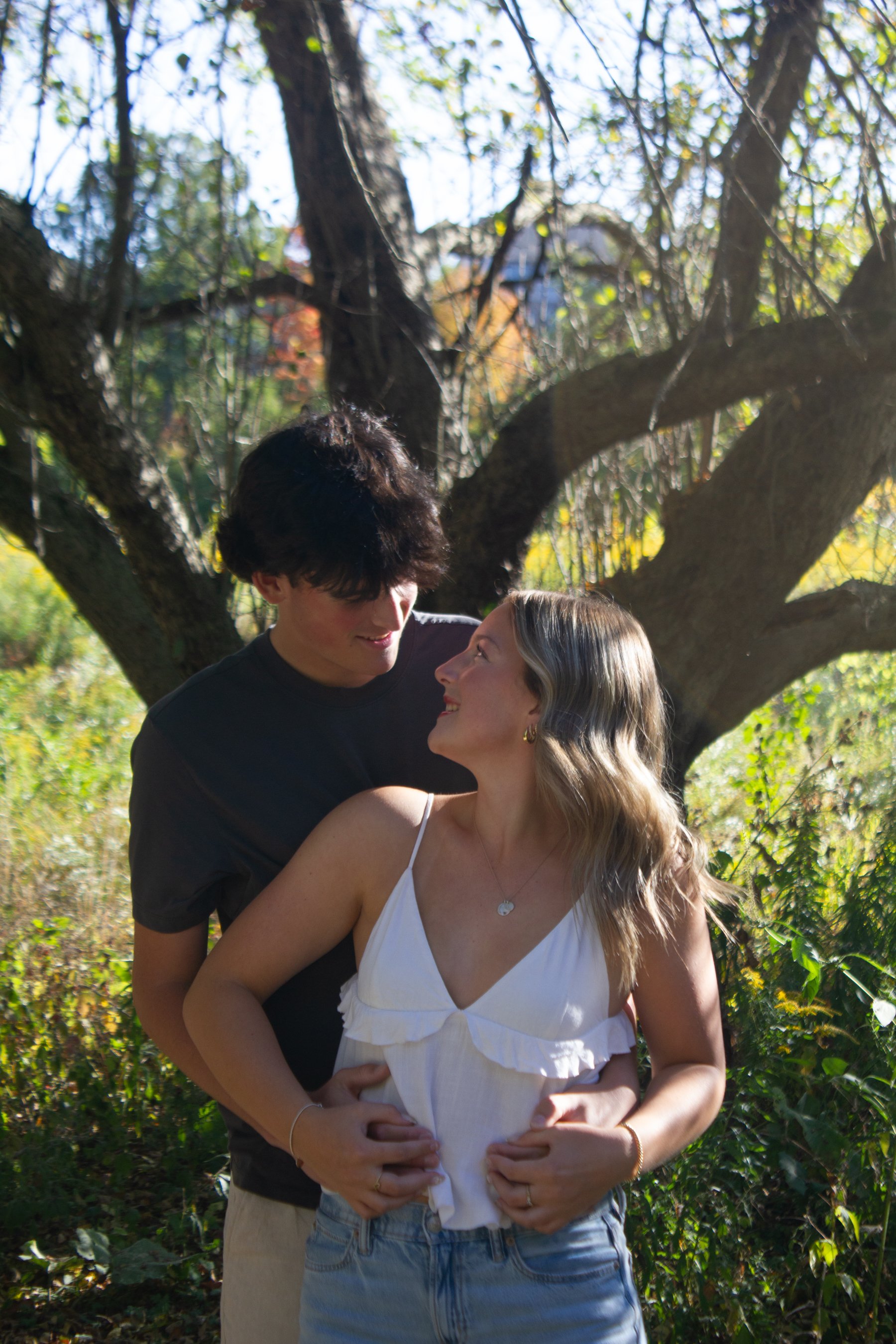 A young couple embraces outdoors in front of a tree, looking into each other's eyes and smiling, with sunlight filtering through the leaves.