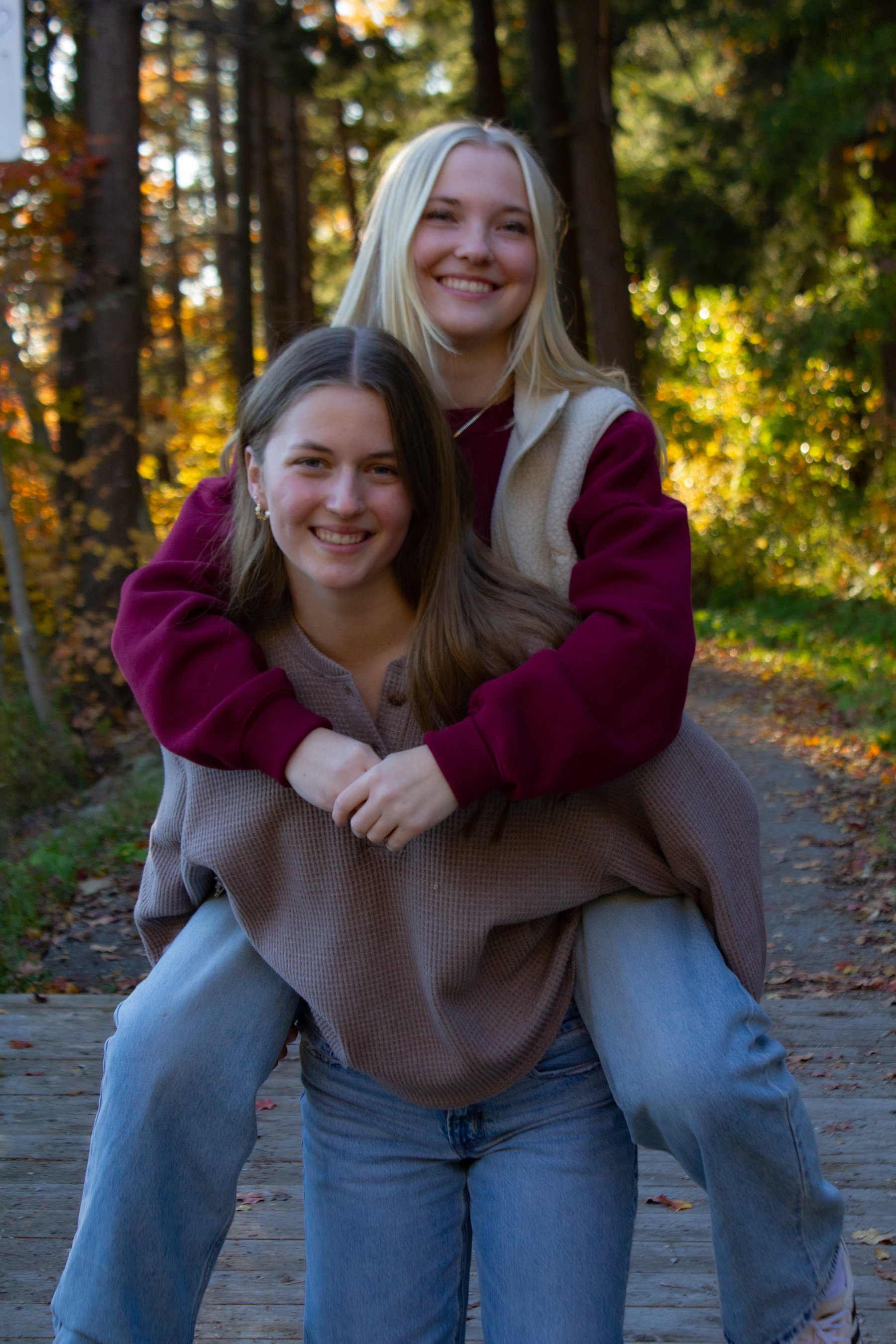 Two young women smiling, one giving a piggyback ride to the other, in a wooded outdoor setting during fall.