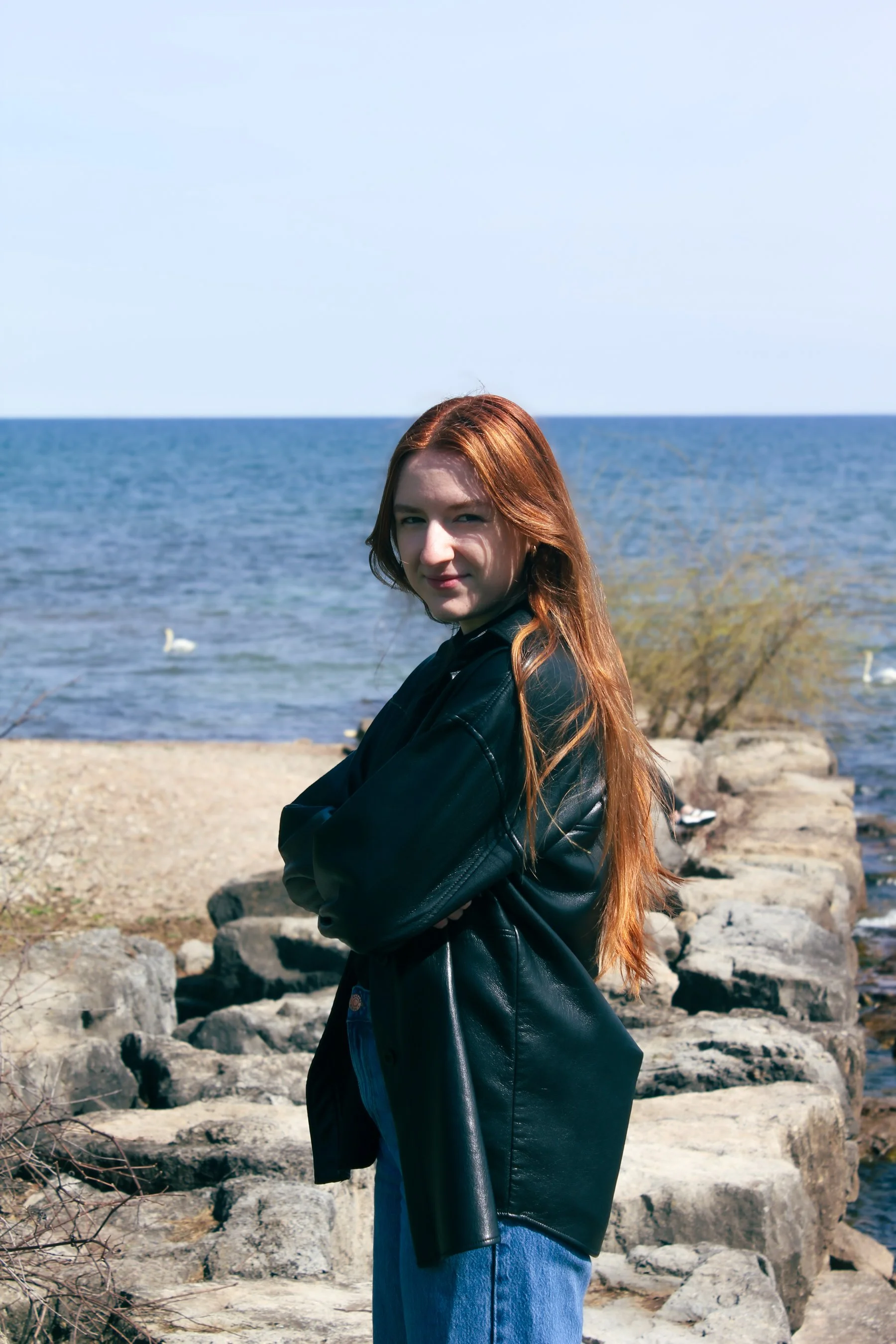 Young woman with long red hair standing on rocky shoreline by the water, wearing a black leather jacket and blue jeans.