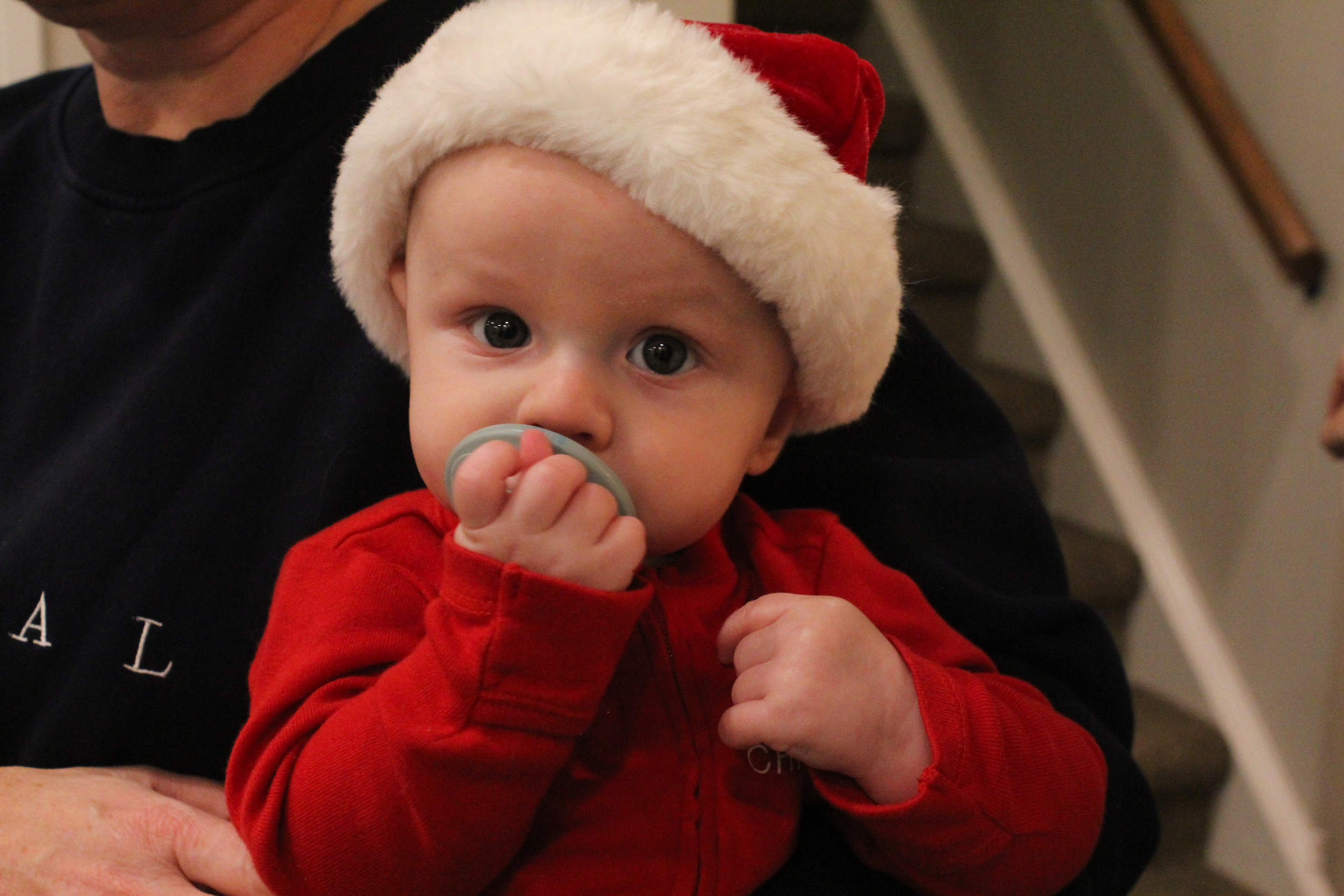 A baby wearing a Santa hat and red jacket, holding a pacifier in their mouth, sitting on someone's lap near a staircase.