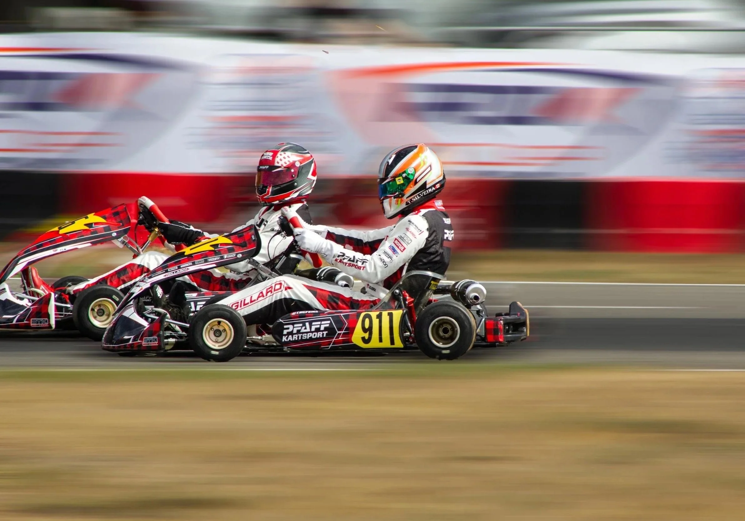 Two go-kart racers competing on a track, with the driver in front wearing a black, white, and red helmet with a white racing suit, and the driver behind wearing a colorful helmet and white racing suit. The go-karts are black and red with numbers and sponsor logos.