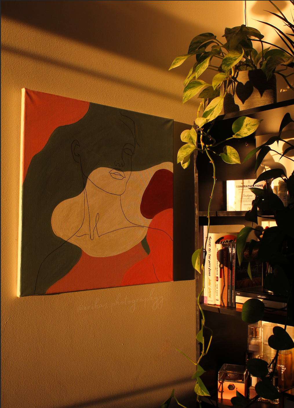 A colorful abstract painting of a woman's face with closed eyes on a beige wall, illuminated by warm lighting, next to a black bookshelf with books, plants, and decorative items.