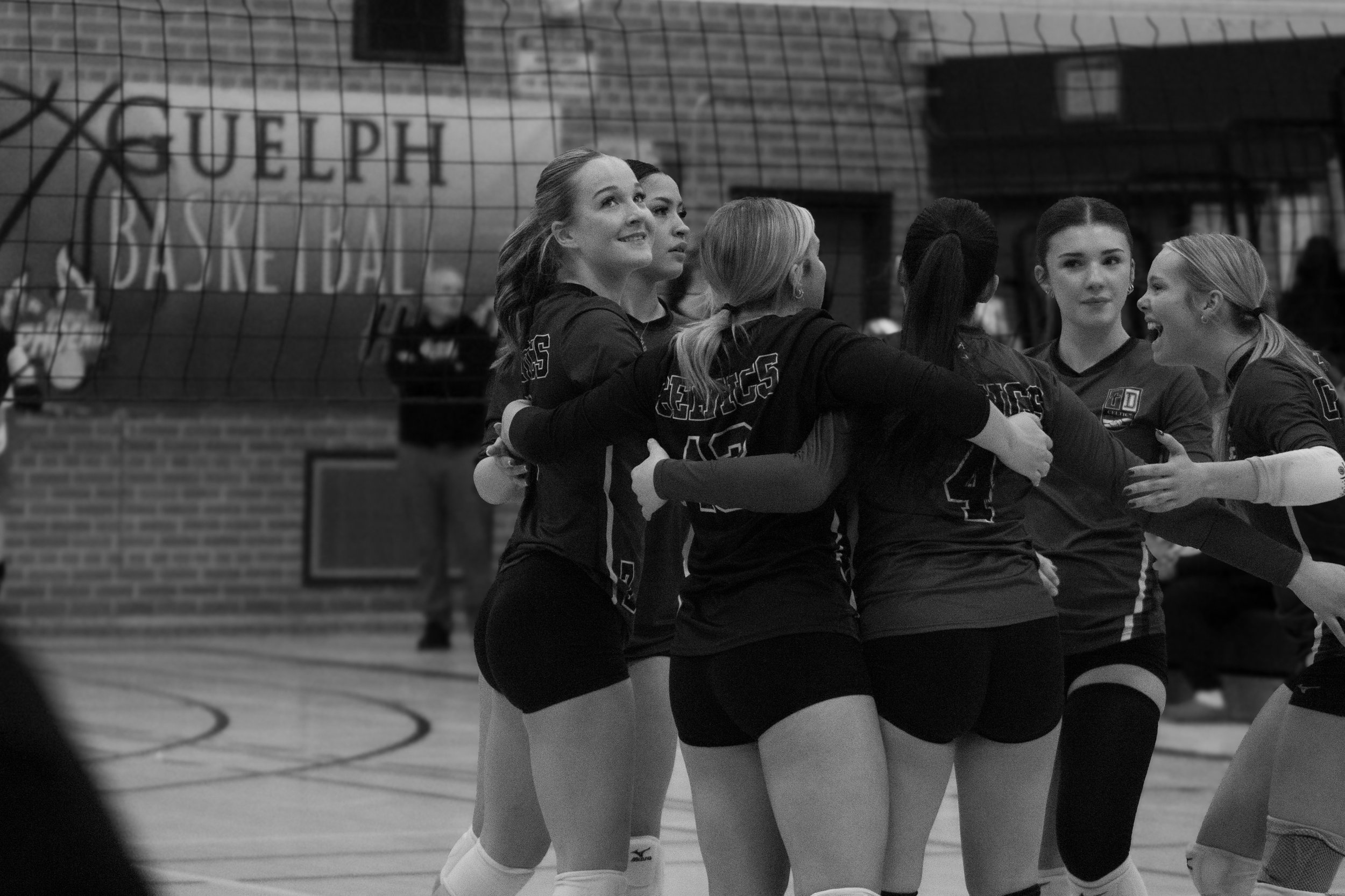 Women volleyball players celebrating together on an indoor court, wearing jerseys with numbers, near a volleyball net.