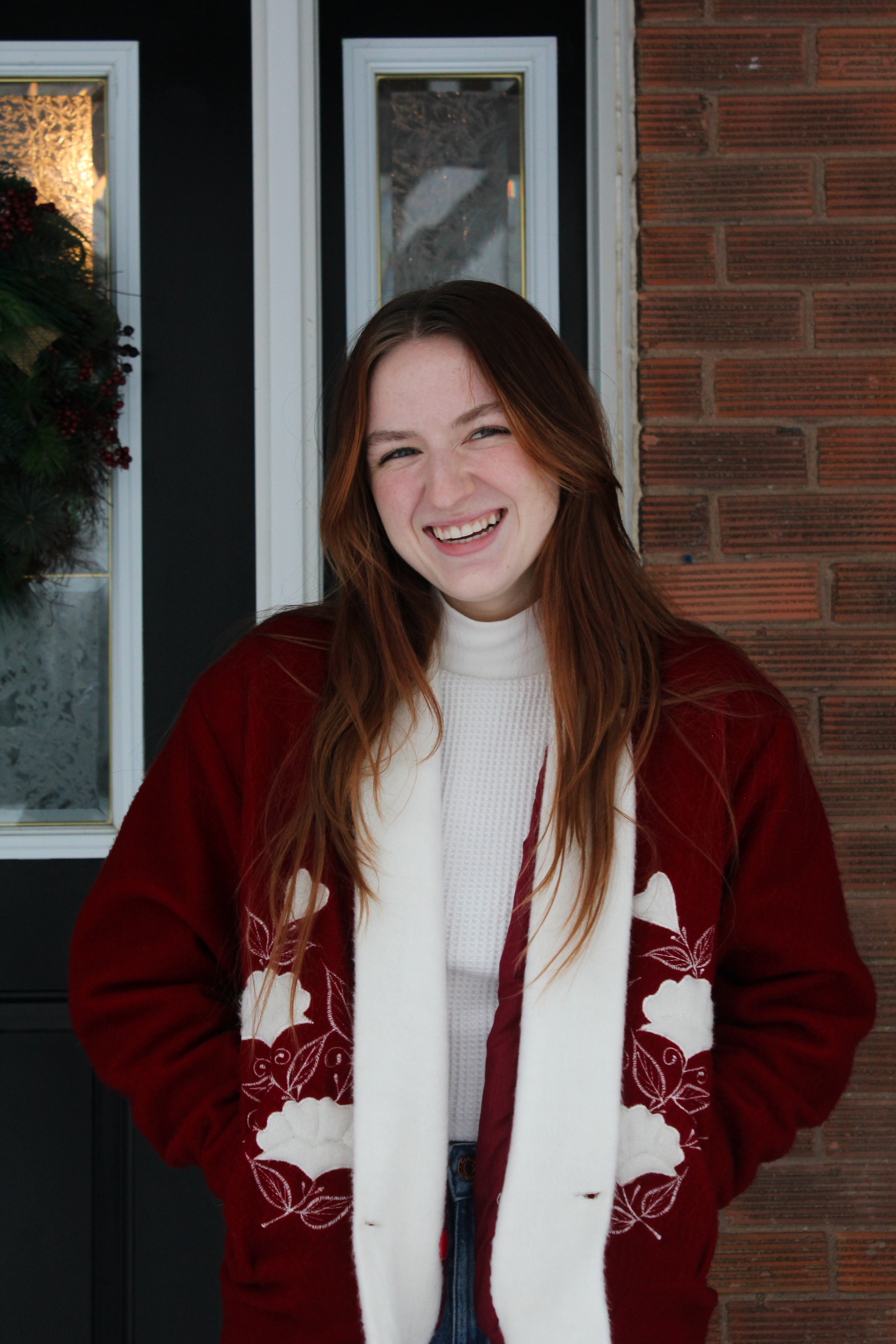 A young woman with long red hair, smiling, standing outside a house with a brick wall and a black door decorated with a holiday wreath.