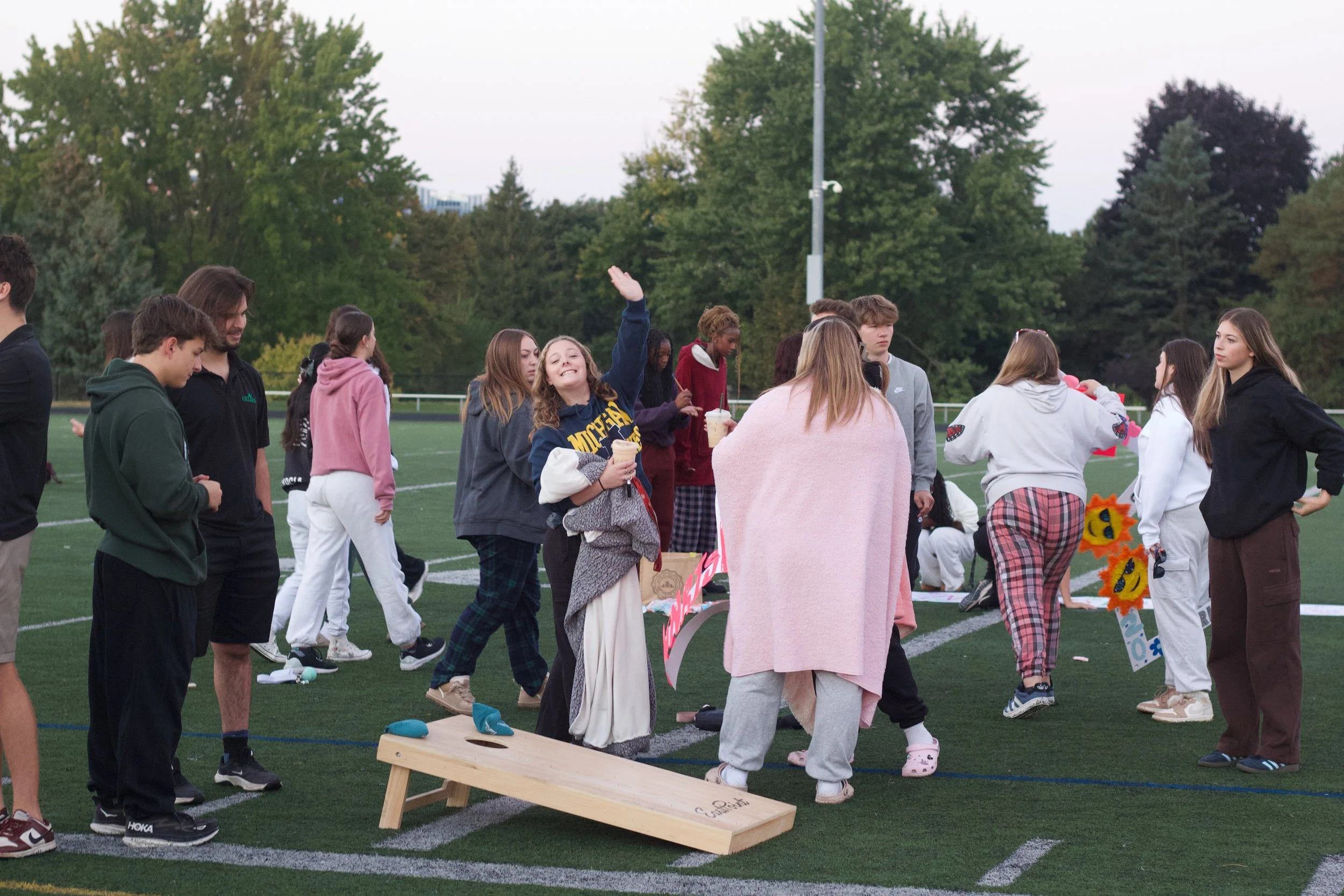 Group of young people gathered on a sports field in the evening, some holding drinks, with trees and a utility pole in the background.