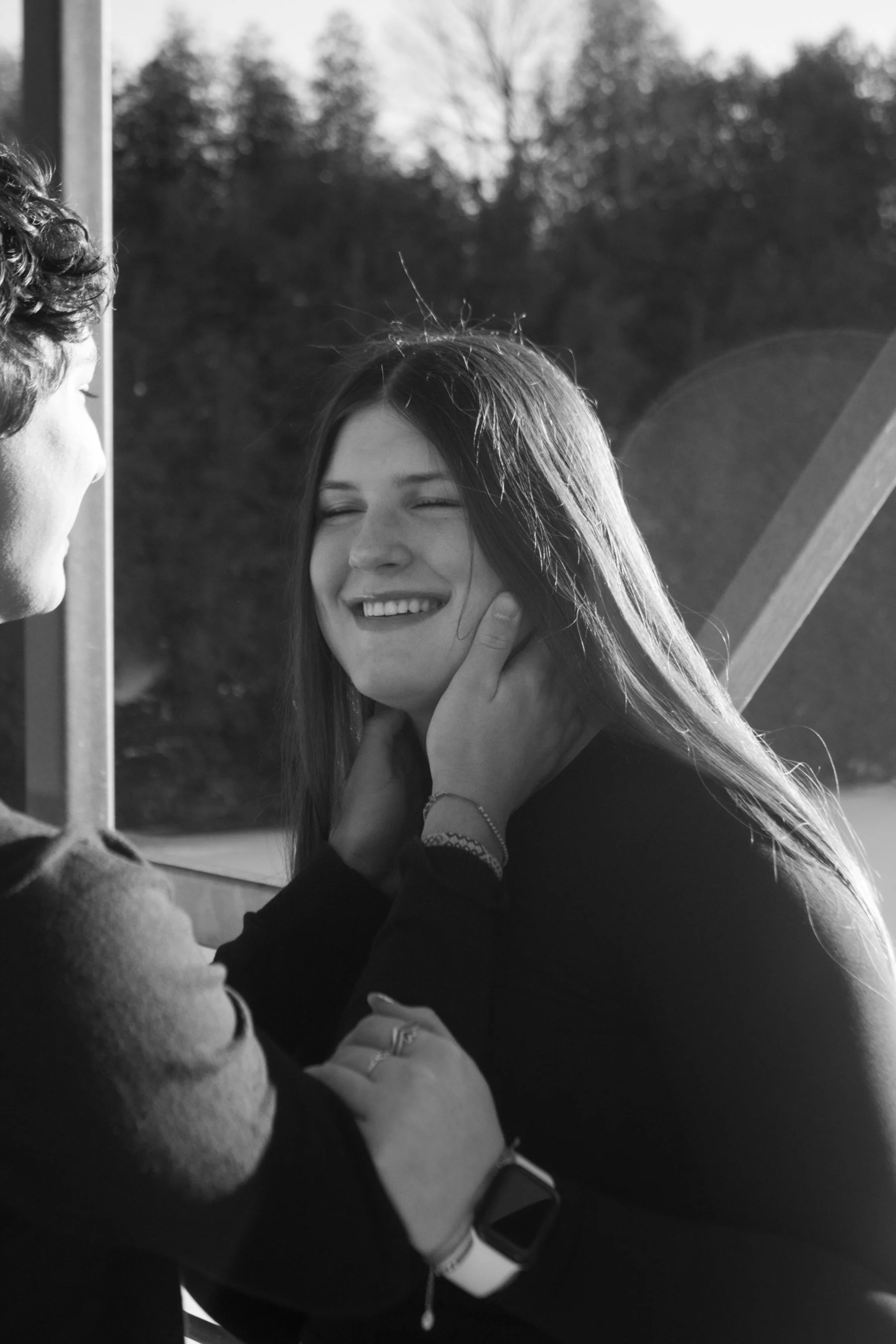 A black and white photo of a young woman with long hair smiling and touching her face, with a man in front of her holding her chin and neck, outdoors with trees in the background.