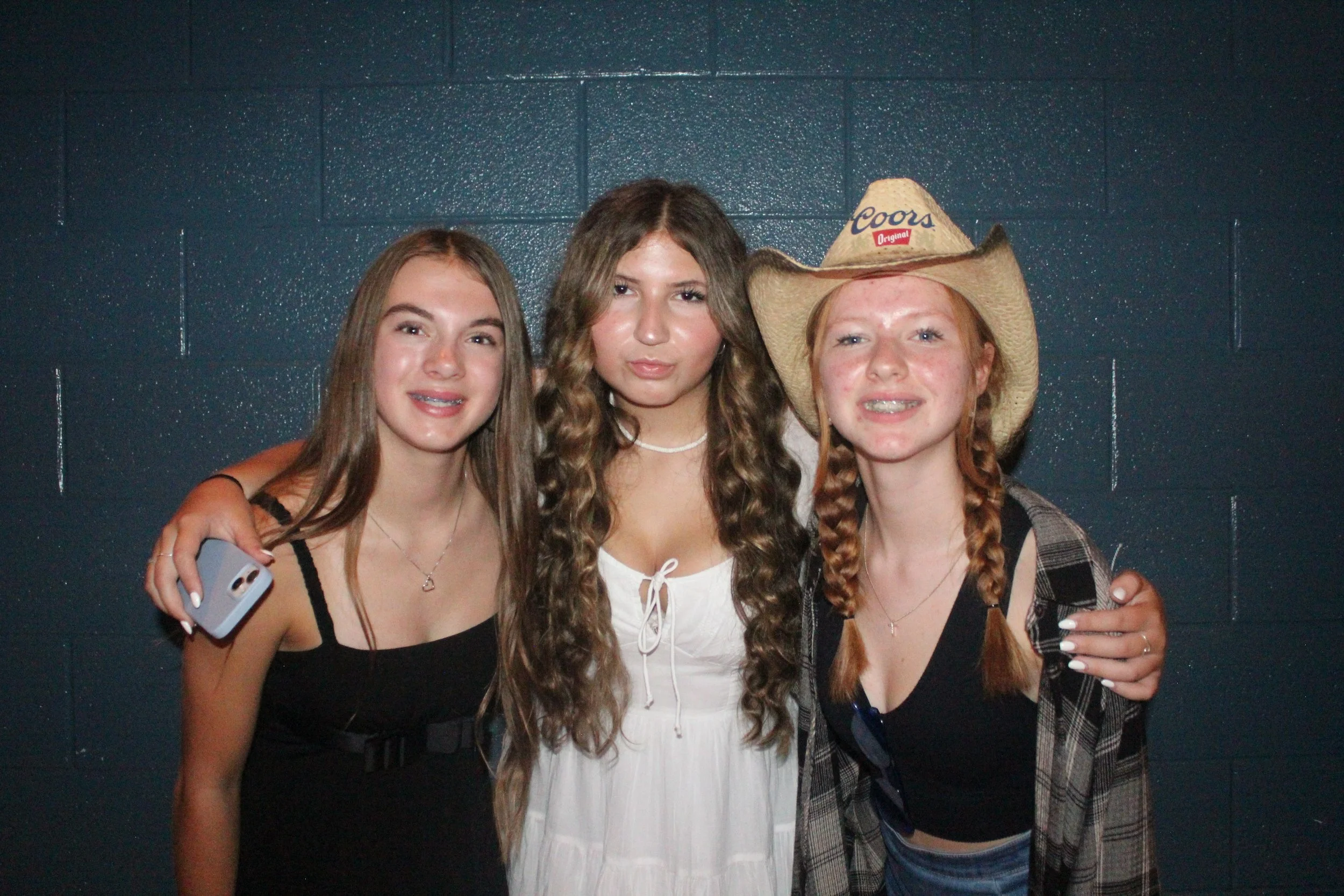 Three young women standing close together against a dark blue tiled wall, smiling and posing for the camera. One is wearing a cowboy hat, while the others have long hair and casual outfits.