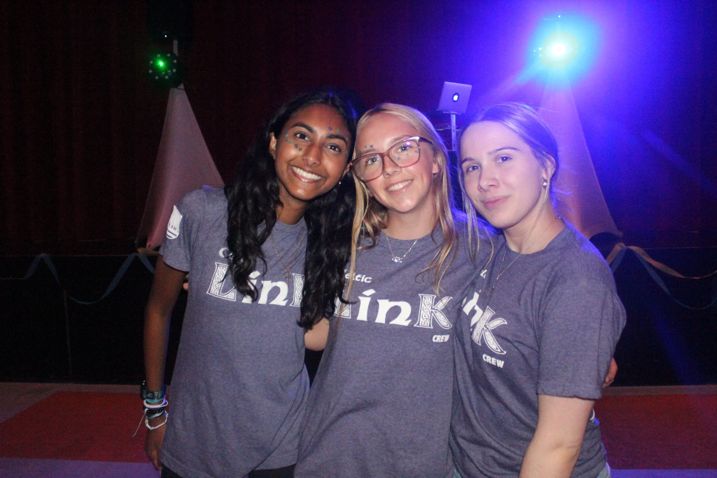 Three young women smiling, wearing matching gray T-shirts that say 'Link Crew', standing close together in front of a dark backdrop with colorful stage lights.