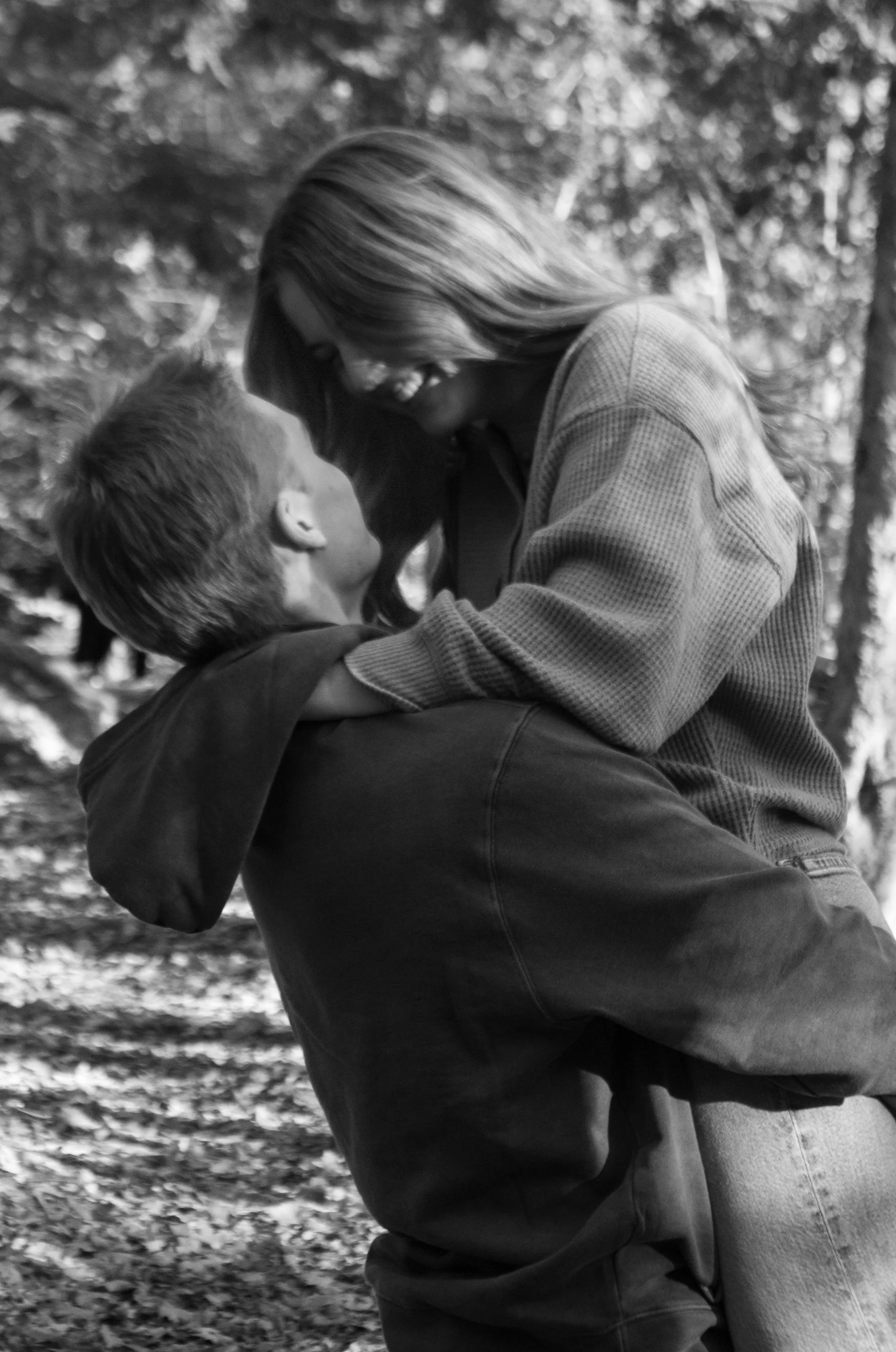A couple sharing a joyful moment outdoors, with the woman sitting on the man's lap, both smiling at each other, surrounded by trees and fallen leaves.