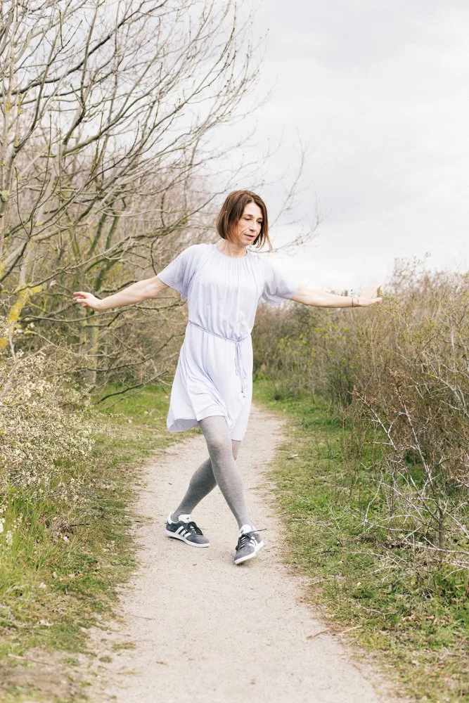 A woman in a white dress and gray tights balancing on a narrow dirt path surrounded by bare trees and bushes.
