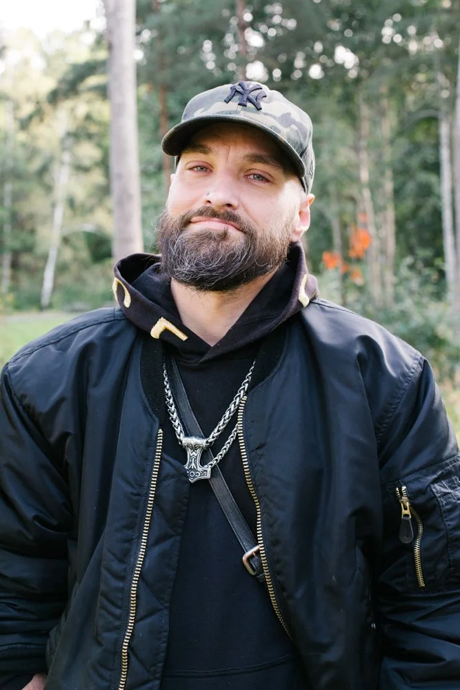 A man with a beard and mustache wearing a camouflage baseball cap, black jacket, black hoodie, and silver chains is standing outdoors in a wooded area with trees in the background.