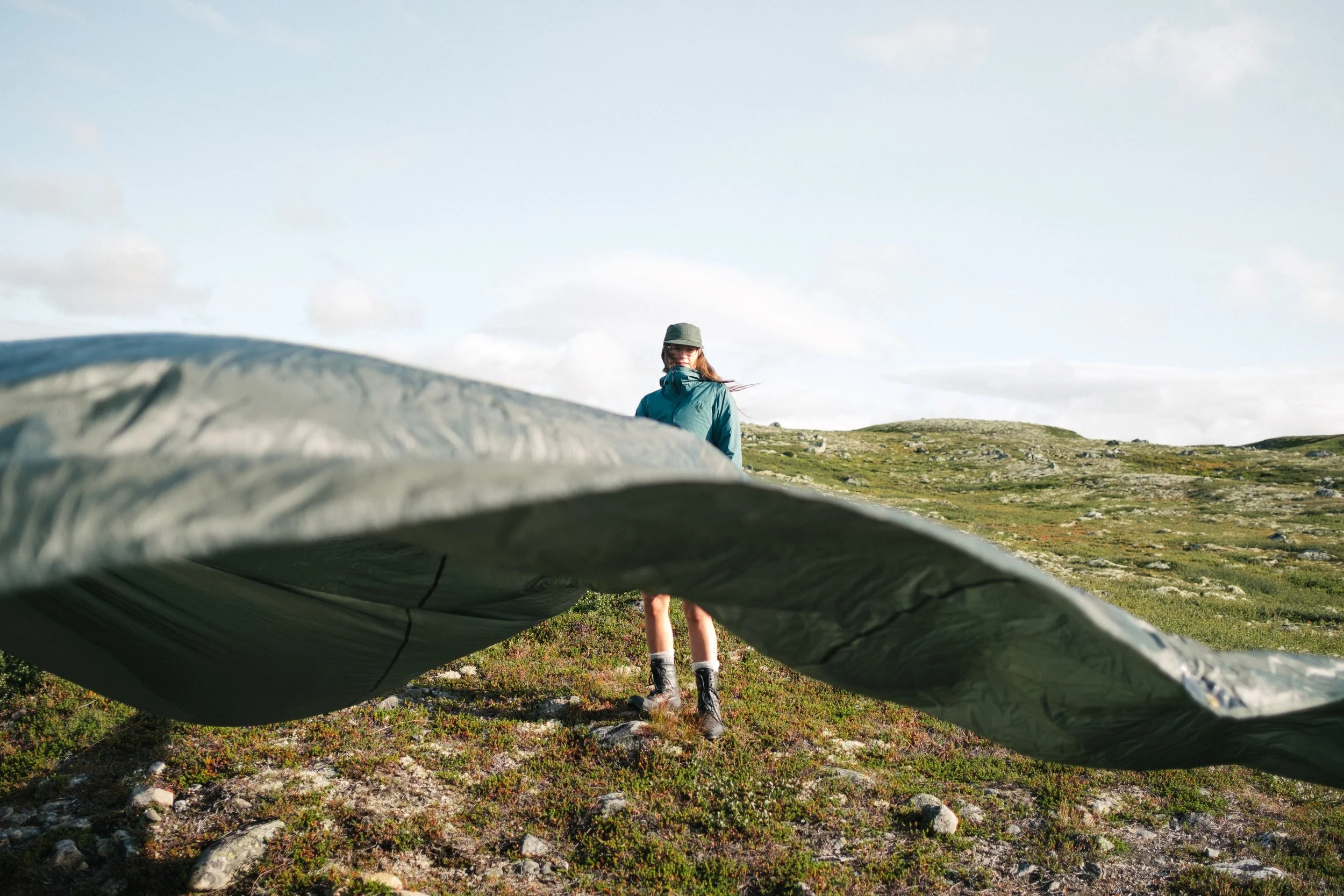 A hiker in a blue jacket and hat stands on a grassy hillside with rocky patches, seen through a large, partially deflated sleeping bag or tarp in the foreground. The background features open landscape with rolling hills and a partly cloudy sky.