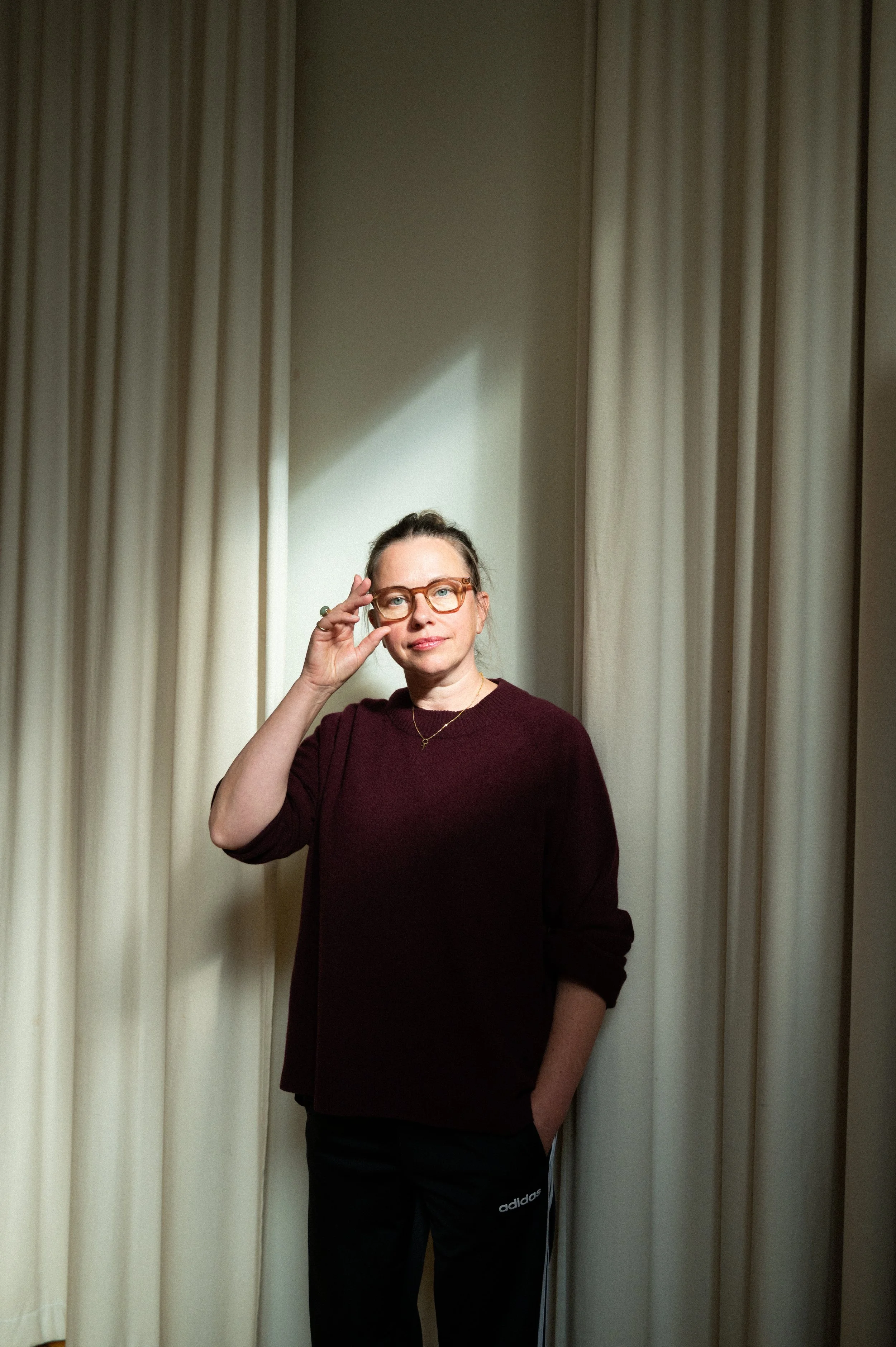 A woman standing in front of cream-colored curtains, wearing glasses, a dark maroon sweater, and black Adidas pants, adjusting her glasses with a hand in her pocket.