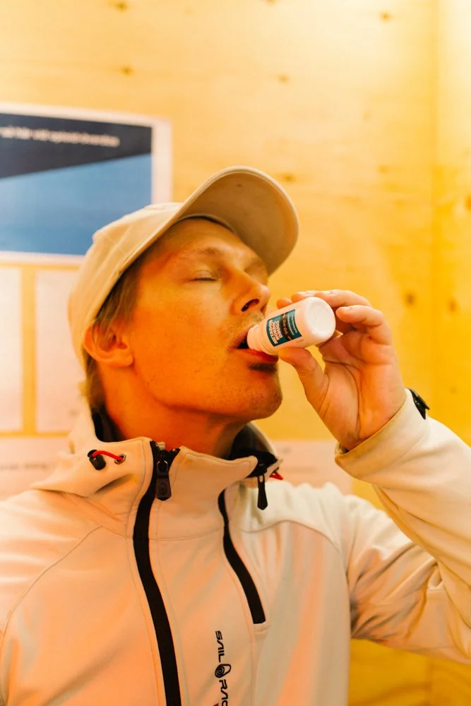 A man in a light-colored jacket and beige cap appears to be taking a pill from a small bottle with his eyes closed.