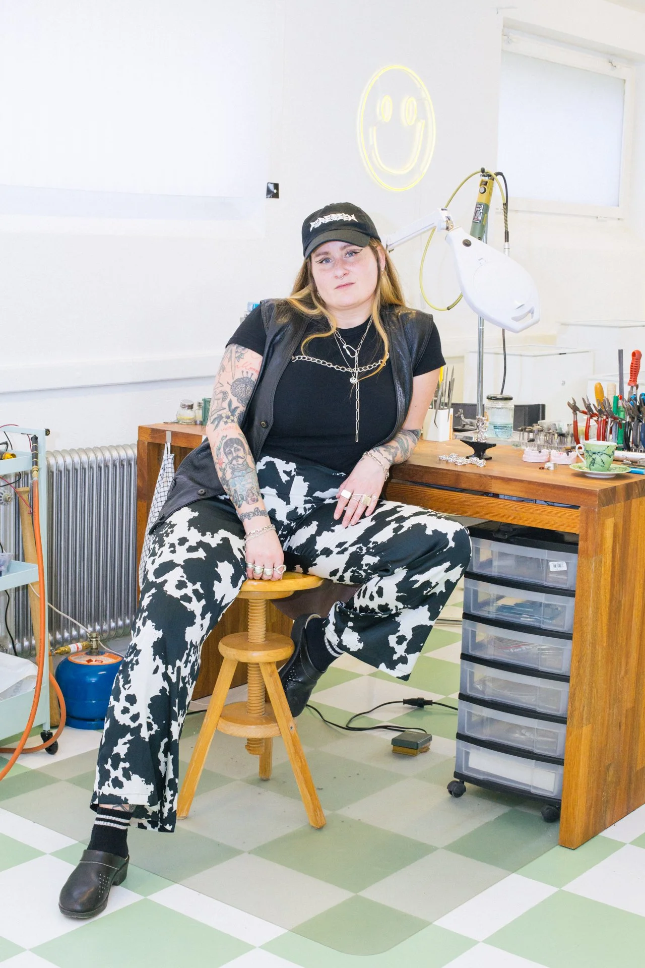 A young woman with tattoos, wearing a black cap, black t-shirt, and cow-print pants, sitting on a wooden stool in a tattoo studio surrounded by tattoo equipment and supplies, with a neon smiley face sign on the wall behind her.