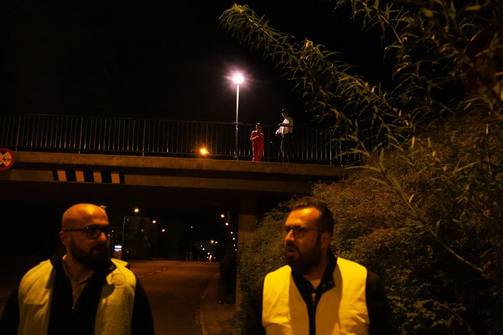 Night scene with two men wearing yellow vests standing on a sidewalk under a bridge, with two people on the bridge in the background near a streetlight.