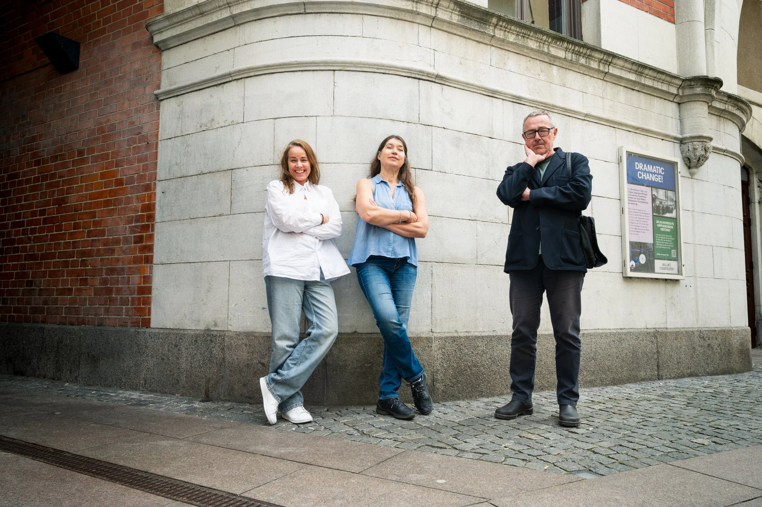 Three people standing on a cobblestone sidewalk in front of a curved building wall. A woman in white with crossed arms, smiling; a woman in a blue sleeveless shirt with crossed arms, looking confident; a man in dark jacket with crossed arms and hand 
