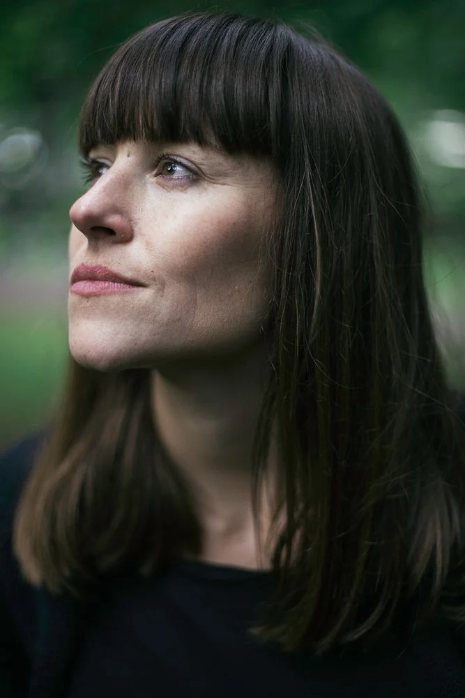 Close-up of a woman with straight dark hair and blunt bangs, looking to the left outdoors with blurred green background.