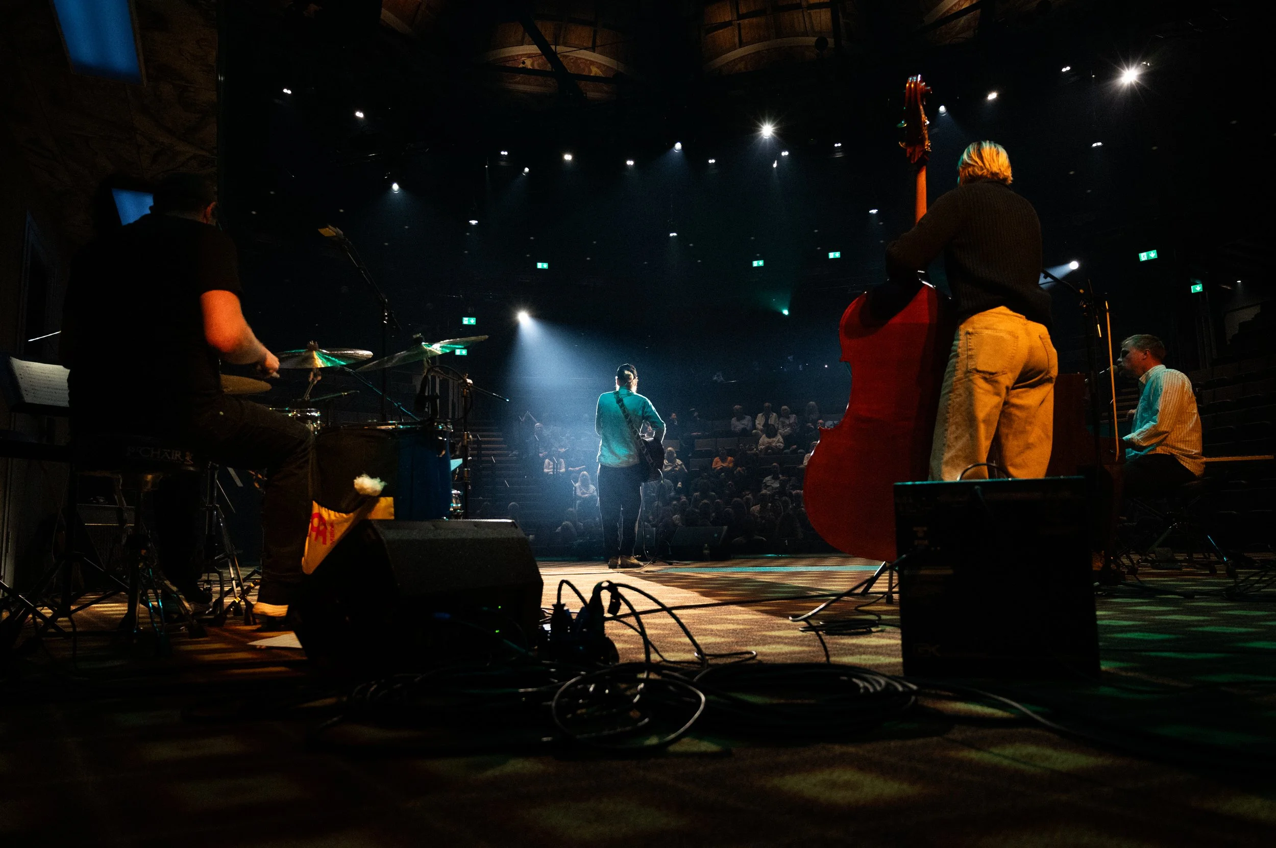 Musicians performing on stage in a dimly lit venue with the audience seated in the background. The band includes a drummer, a bassist, and a keyboardist, with a performer in the center holding a guitar.