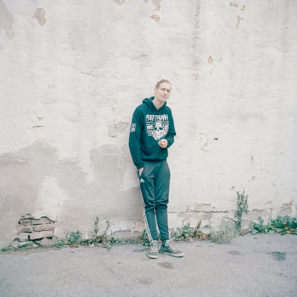 A young man in a teal hoodie and gray jogger pants stands against a weathered, off-white brick wall with peeling paint and some small plants at the base.