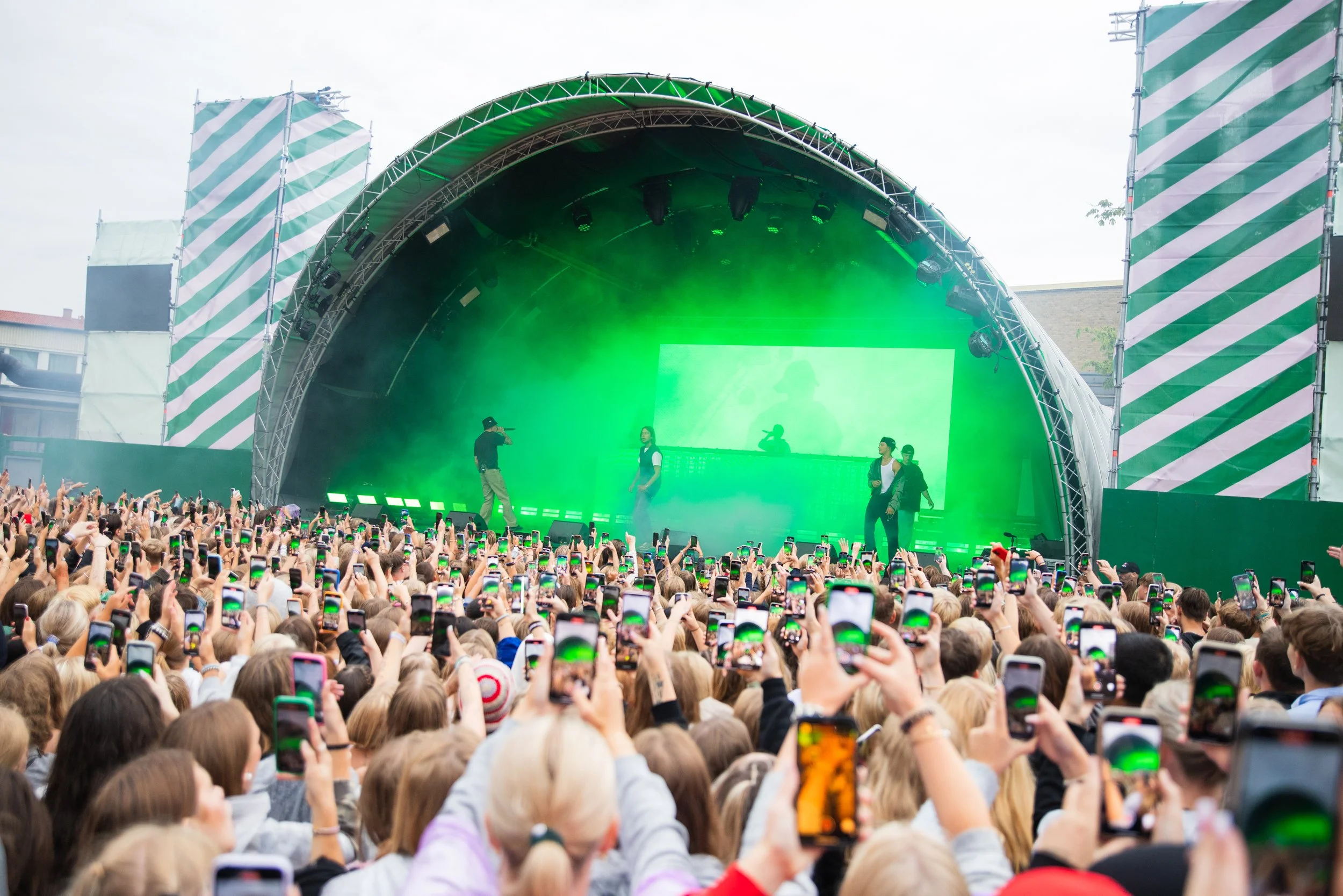 Large outdoor concert with a crowd holding up smartphones, a stage with three performers, green lighting, and a large screen in the background.