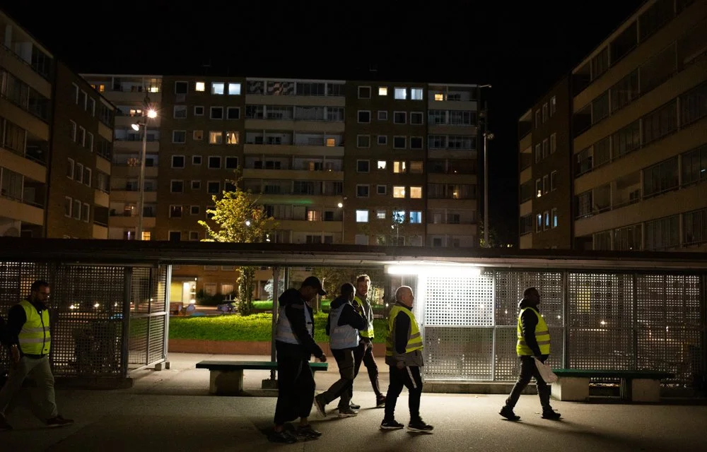 A group of people wearing yellow reflective vests walk through a city area at night, near a bus stop or shelter with benches, surrounded by tall apartment buildings with lit windows.