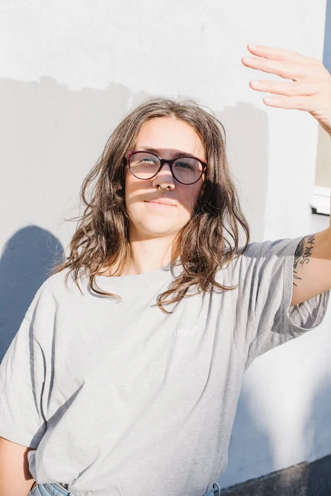 Young woman with glasses, curly brown hair, and a septum piercing, wearing a light gray T-shirt, shielding her eyes from the sun with her hand, standing against a white wall.