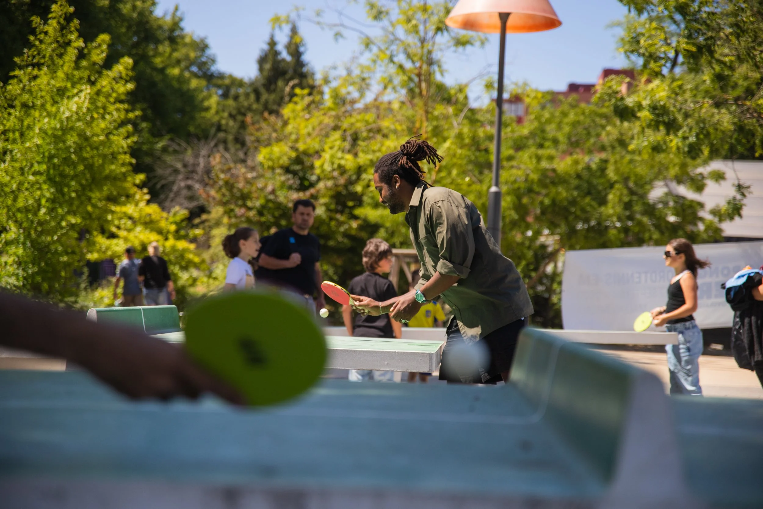 People playing table tennis outdoors on a sunny day, with trees and a lamp post in the background.