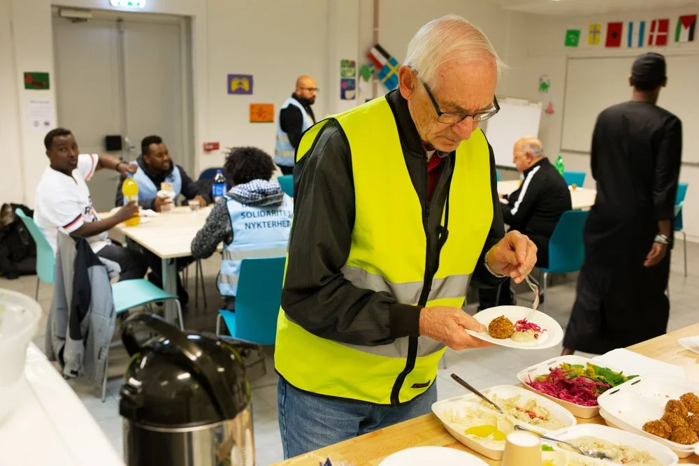 An elderly man in a yellow safety vest is serving himself food from a buffet table at a community event. In the background, a group of people is sitting at a table, some with drinks, and colorful international flags decorate the wall.