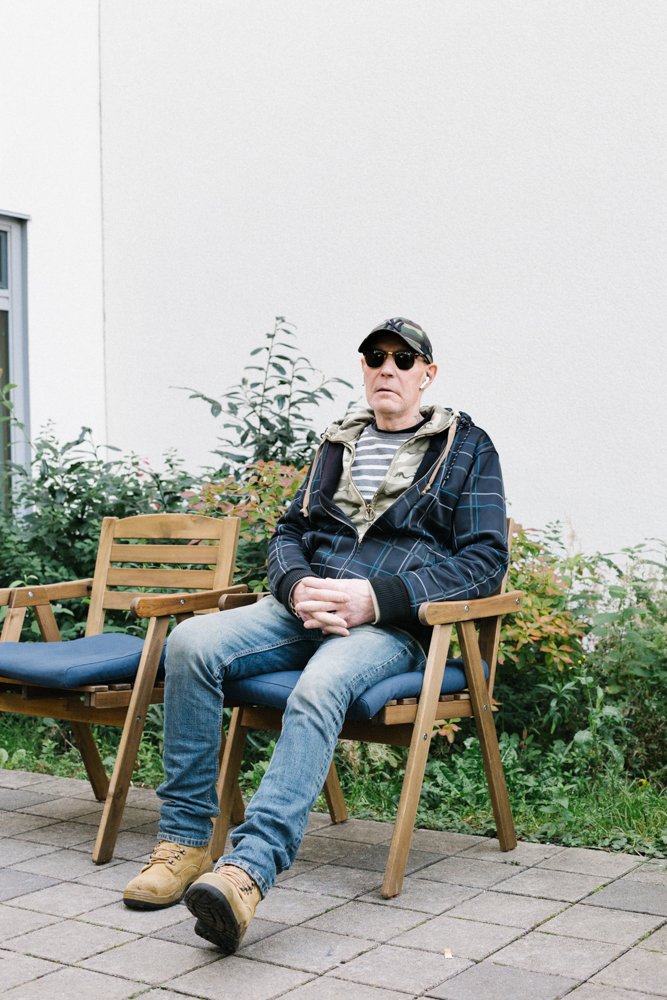 An older man wearing sunglasses, a camouflage hat, a striped t-shirt, a hoodie, and a jacket, sitting outdoors on a wooden bench with a cushion, surrounded by greenery and a white wall.