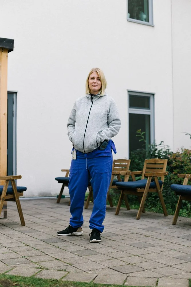 A woman standing outdoors on a paved area, wearing a gray zip-up jacket, blue pants, and black sneakers, with wooden chairs arranged around her.