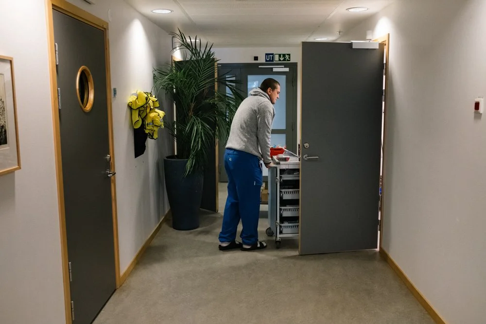 A man in a gray hoodie and blue pants standing in a hallway next to a mobile medical cart, with a notepad and medical supplies on top. There is a large potted plant nearby and two closed doors, one with a circular window. The hallway has cream walls,