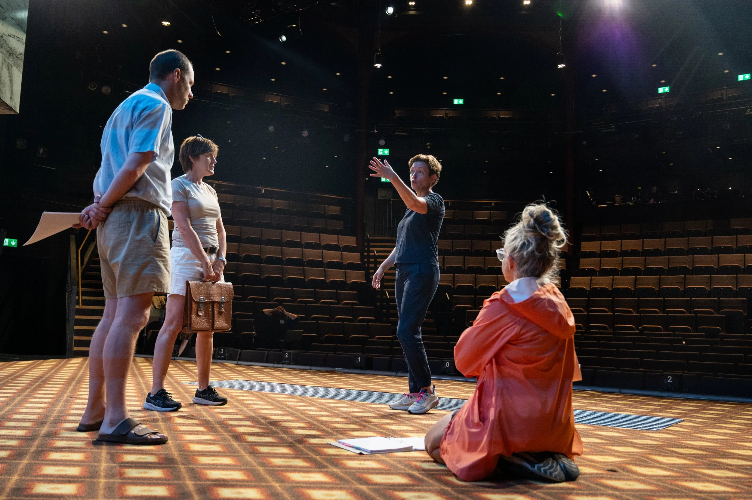 A group of four people on a stage in a theatre, with two men and two women, appears to be rehearsing or discussing a performance. One woman is sitting on the ground, while the others are standing, and one person is gesturing with an arm raised.