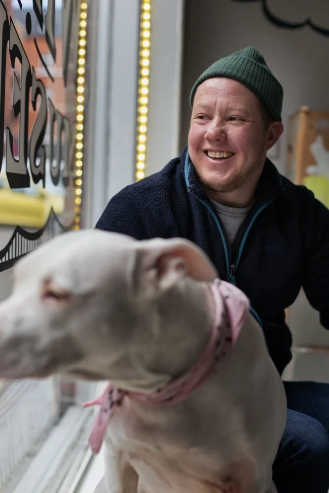 A smiling man wearing a green beanie and navy fleece hoodie sitting next to a white pitbull with a pink collar, indoors near a window with lights in the background.