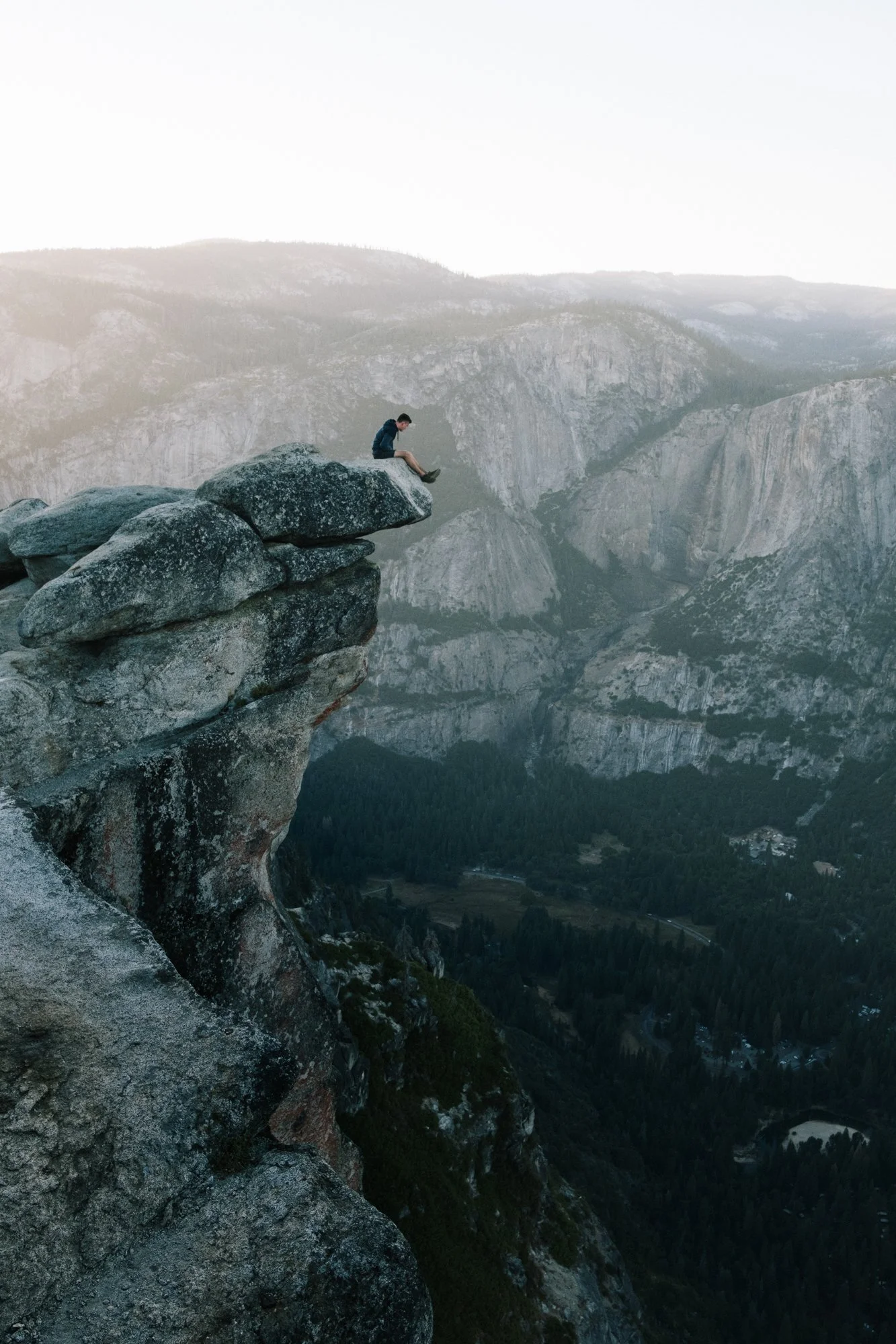 A person sitting on the edge of a large rock formation overlooking a mountainous landscape.