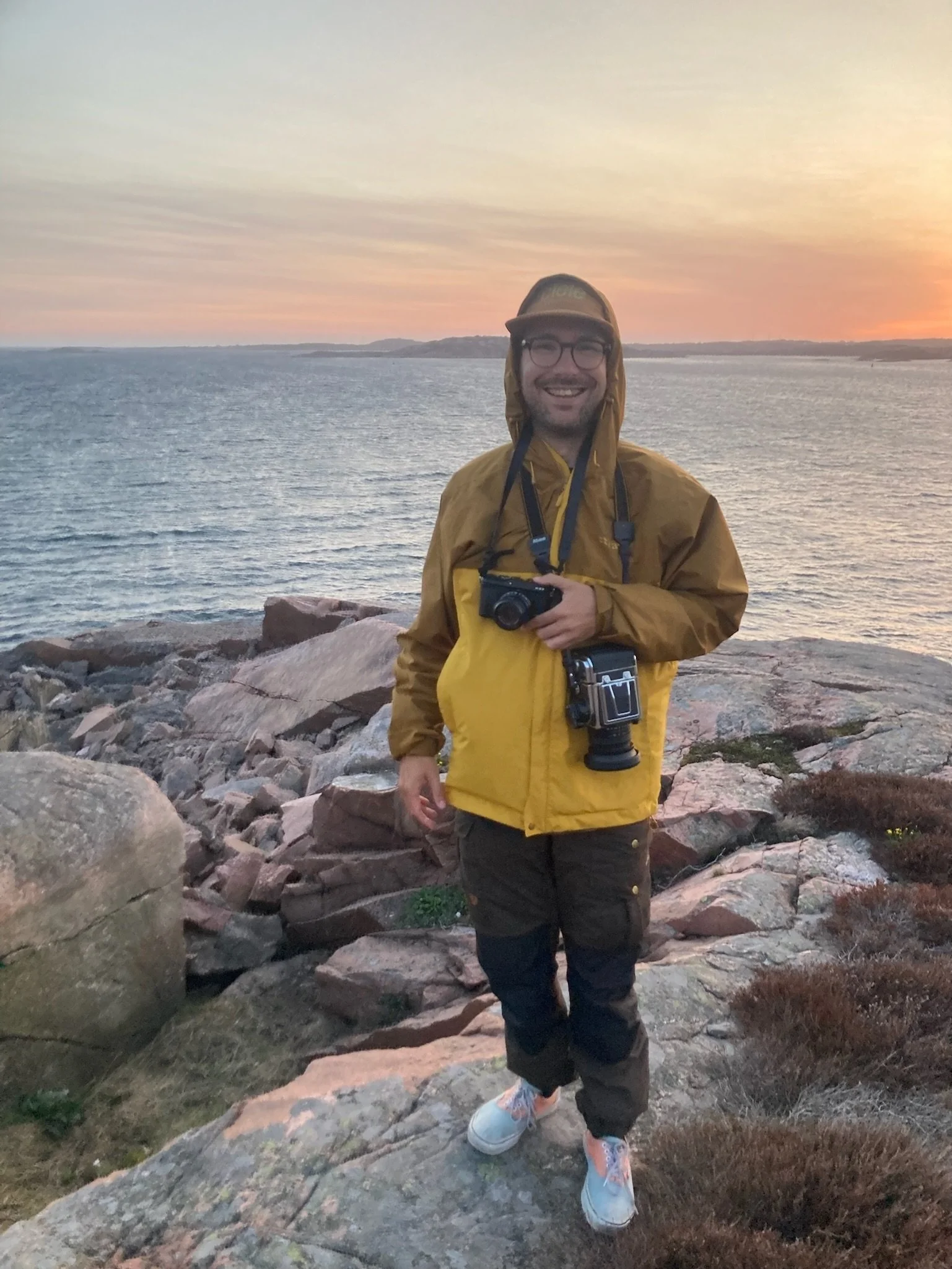 Picture of Mikael on a cliff, ocean and sunset in the background. He carries two cameras around his neck and looks happy.
