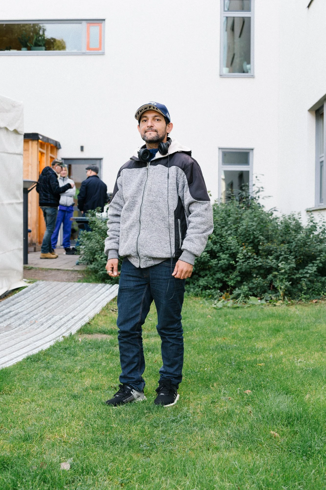 A man standing on a grassy yard, wearing a grey hoodie jacket, jeans, and black sneakers, with headphones around his neck, and a group of people in the background near a modern white building.