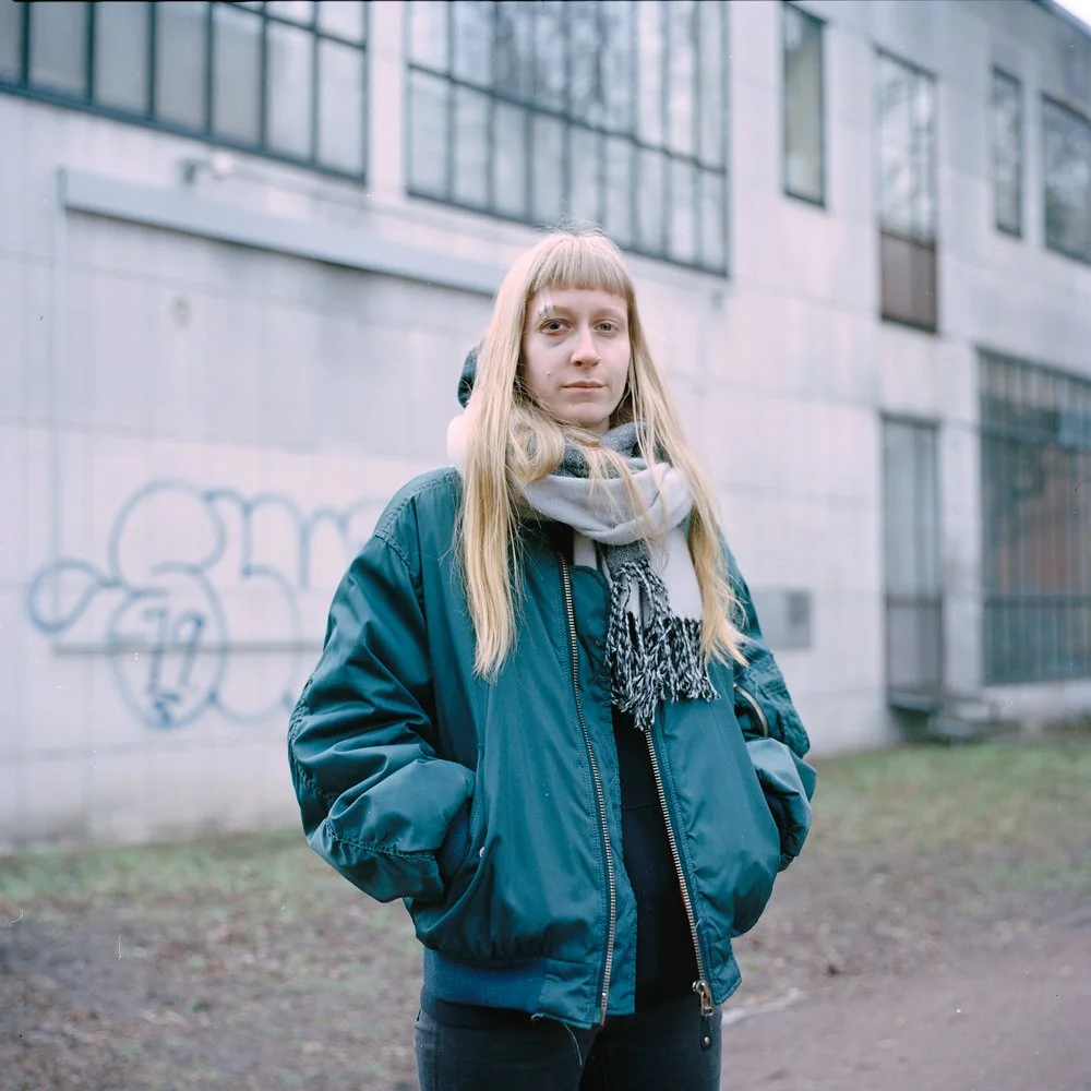 A young woman with long blonde hair and bangs wearing a dark blue jacket and a gray scarf, standing outdoors in front of a concrete building with graffiti on the wall.