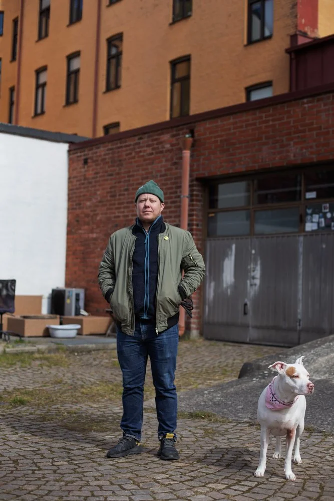 A man wearing a green beanie, olive jacket, and blue jeans standing outdoors with his hands in his jacket pockets, next to a white dog wearing a pink bandana, in an urban setting with brick buildings.