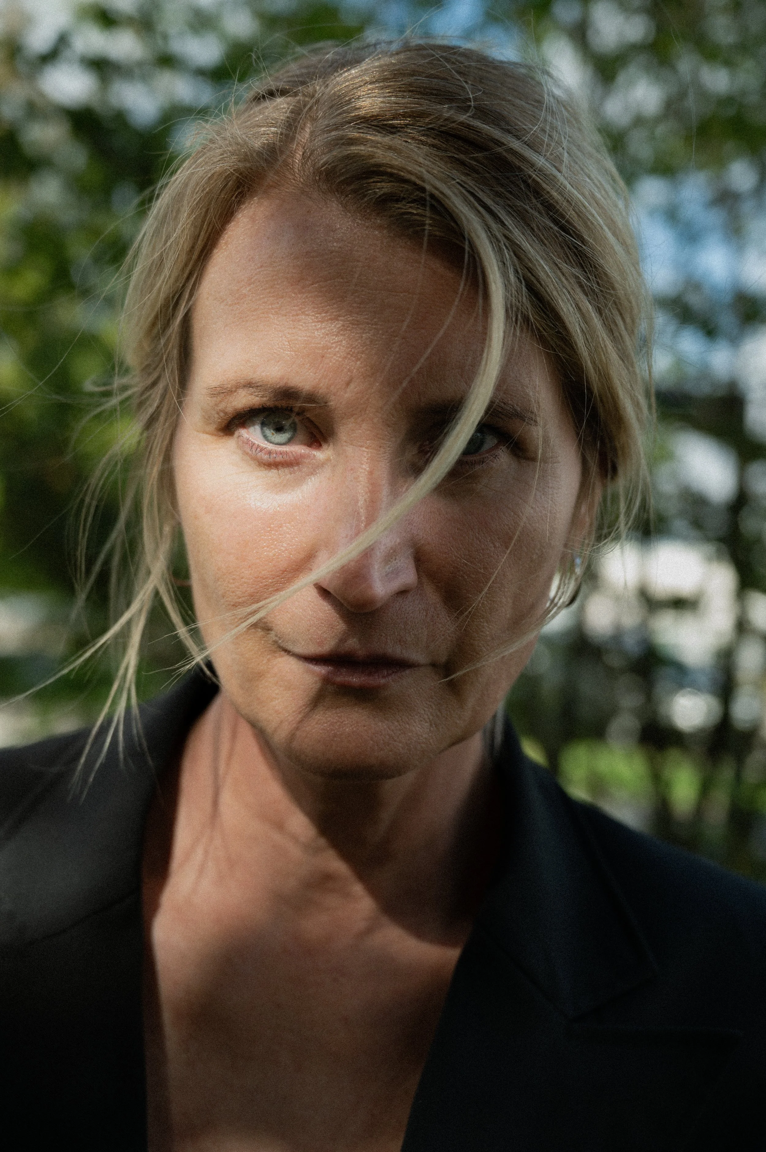 Close-up portrait of a woman with blonde hair and blue eyes, outdoors with blurred green trees in the background.